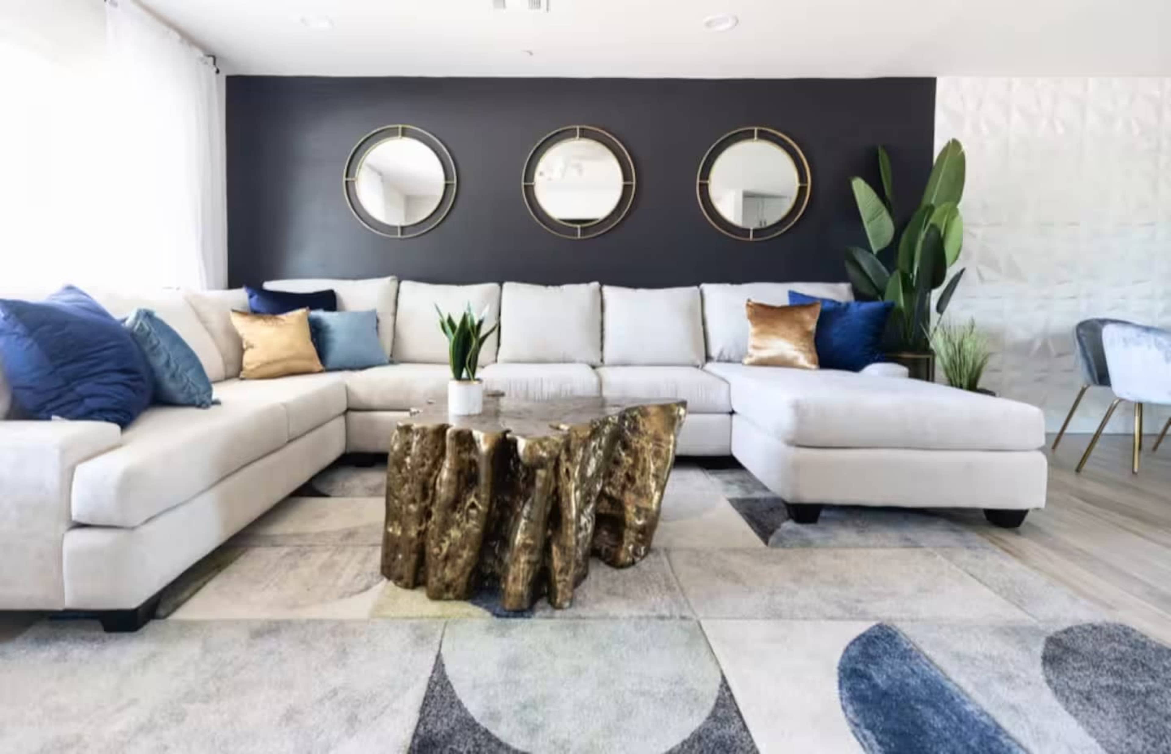 A modern living room features a large beige sectional sofa with decorative pillows, a gold and wood accent table, and three round mirrors on the dark wall, alongside a plant and a patterned rug.