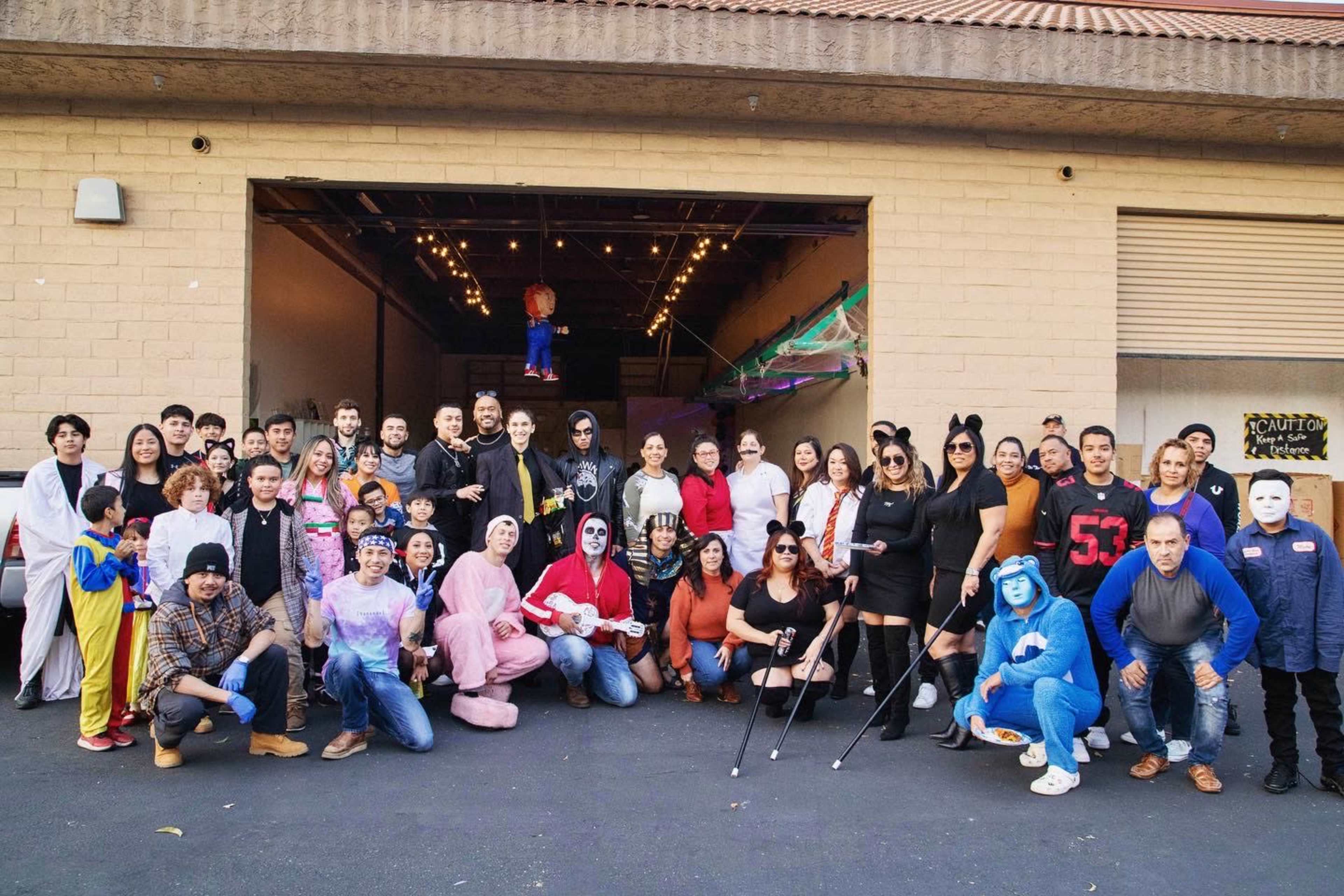 A large group of people poses together in front of a building, many dressed in various costumes and masks for a themed event.