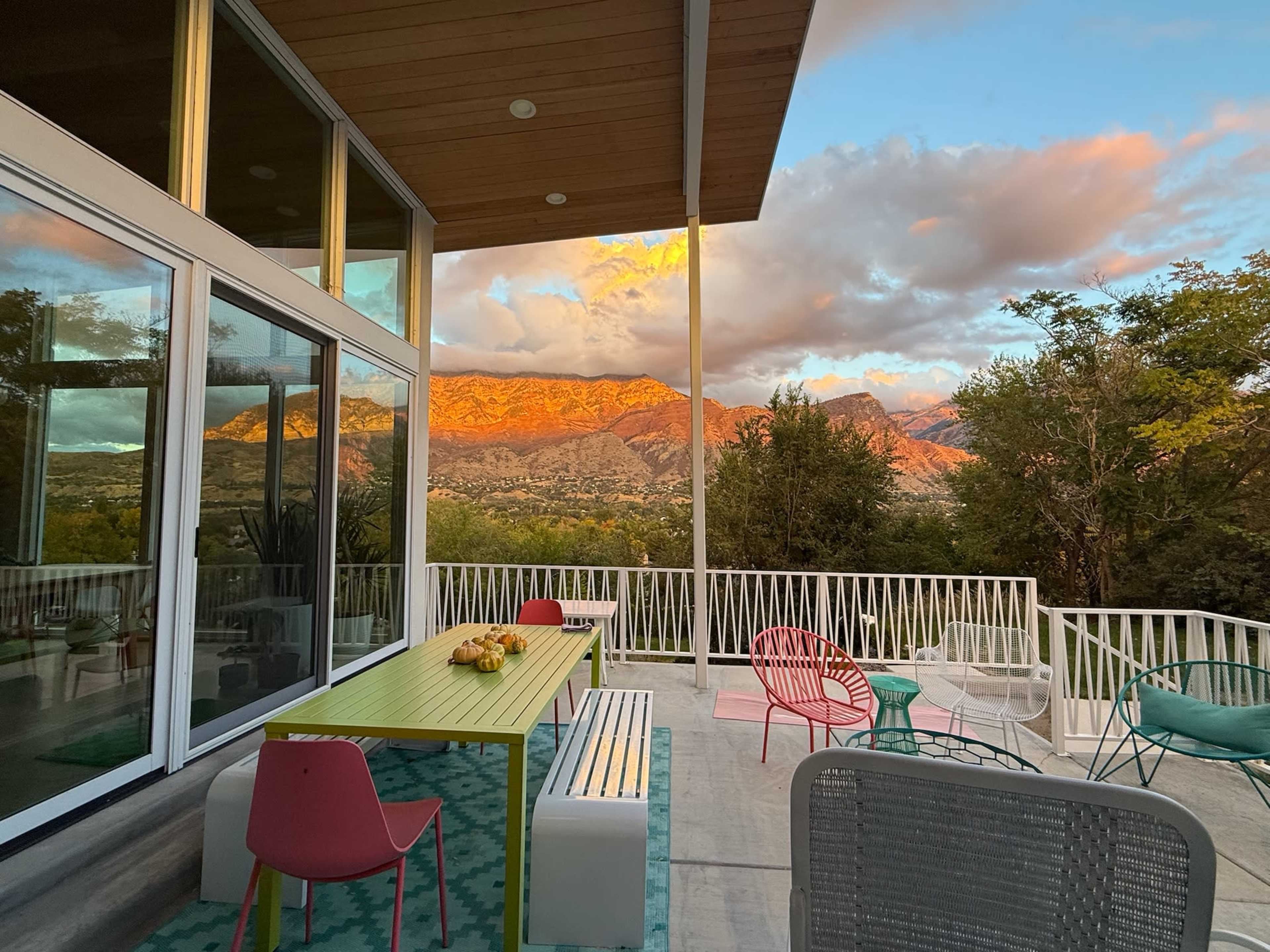 A modern outdoor patio with colorful furniture overlooks a mountainous landscape under a partially cloudy sky at sunset.