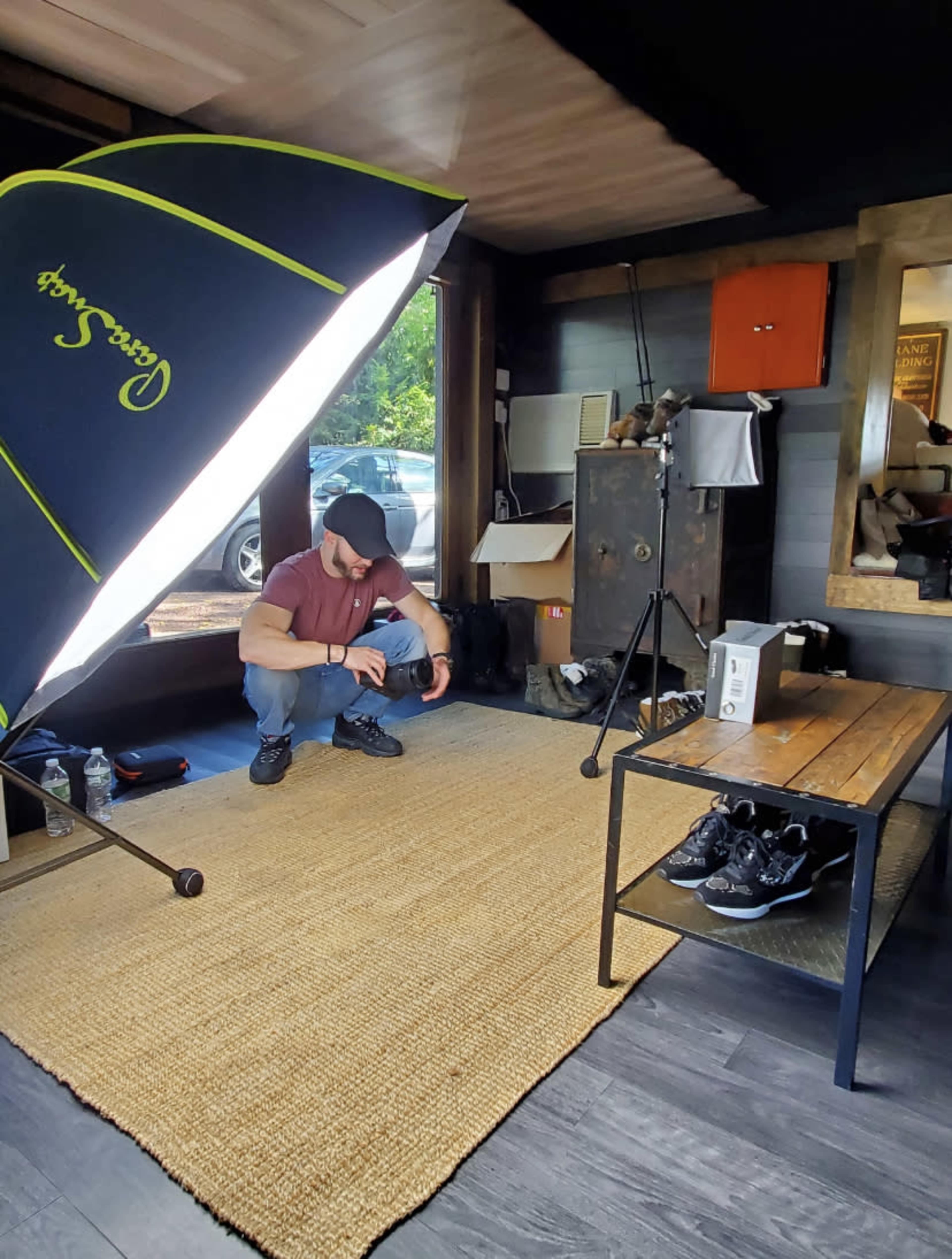 A person is crouching on a rug in a room with a large softbox light and a wooden table displaying shoes, while boxes and a safe are visible in the background.
