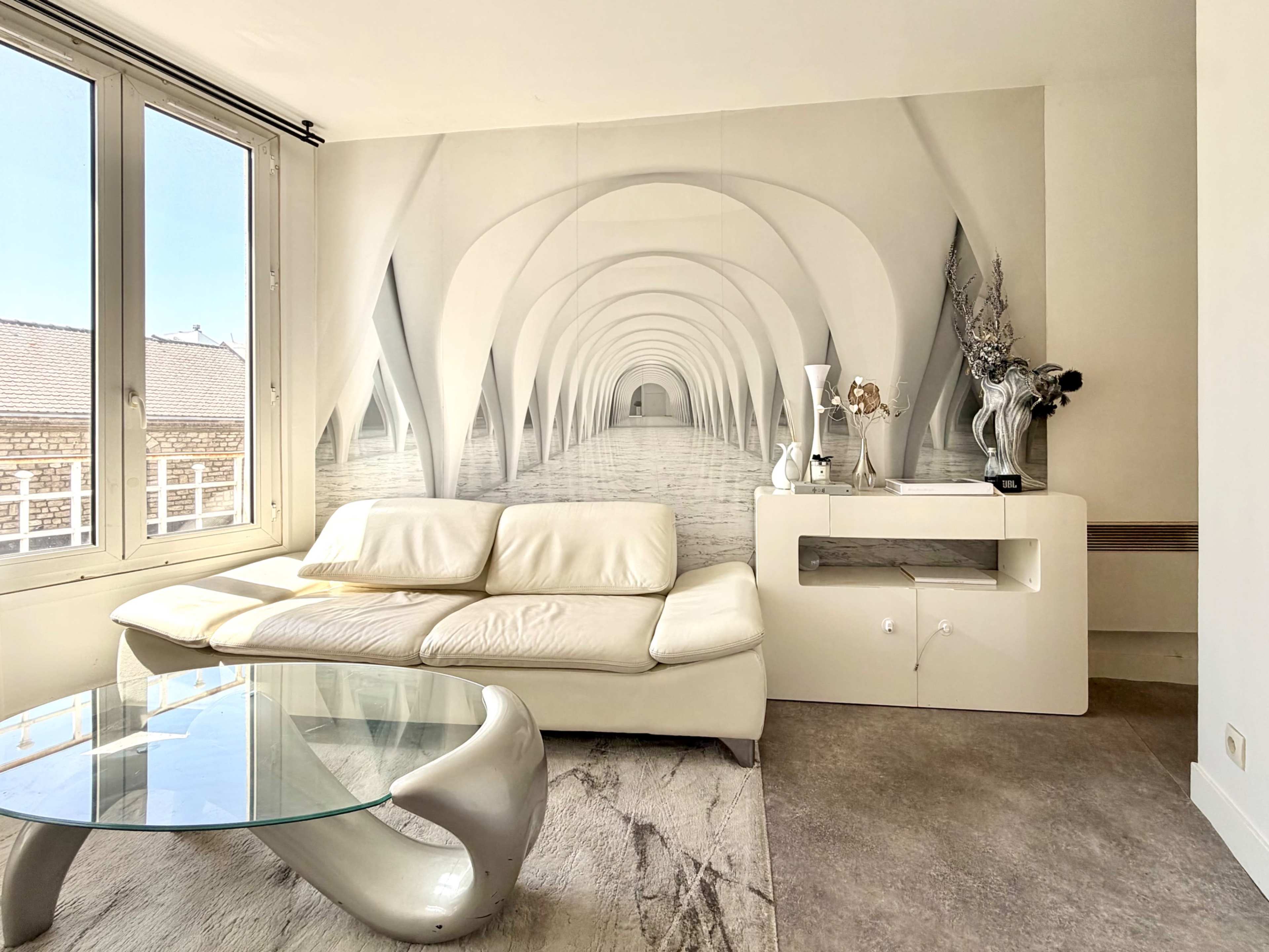 A modern living room features a white leather sofa, a glass coffee table, and a large mural depicting an architectural interior with arches.