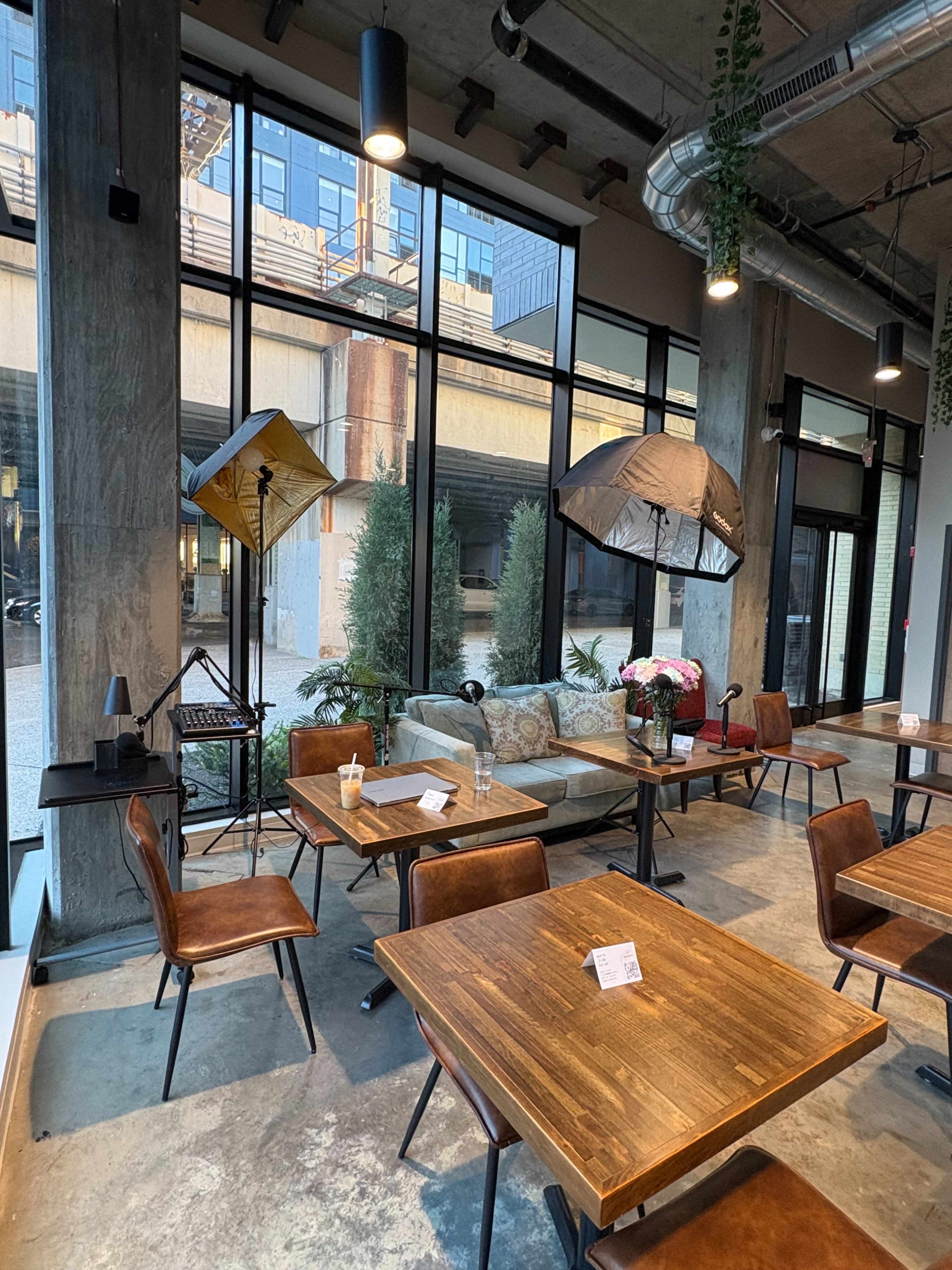The image shows a modern café interior with wooden tables and chairs, large windows letting in natural light, and photography equipment set up near potted plants.