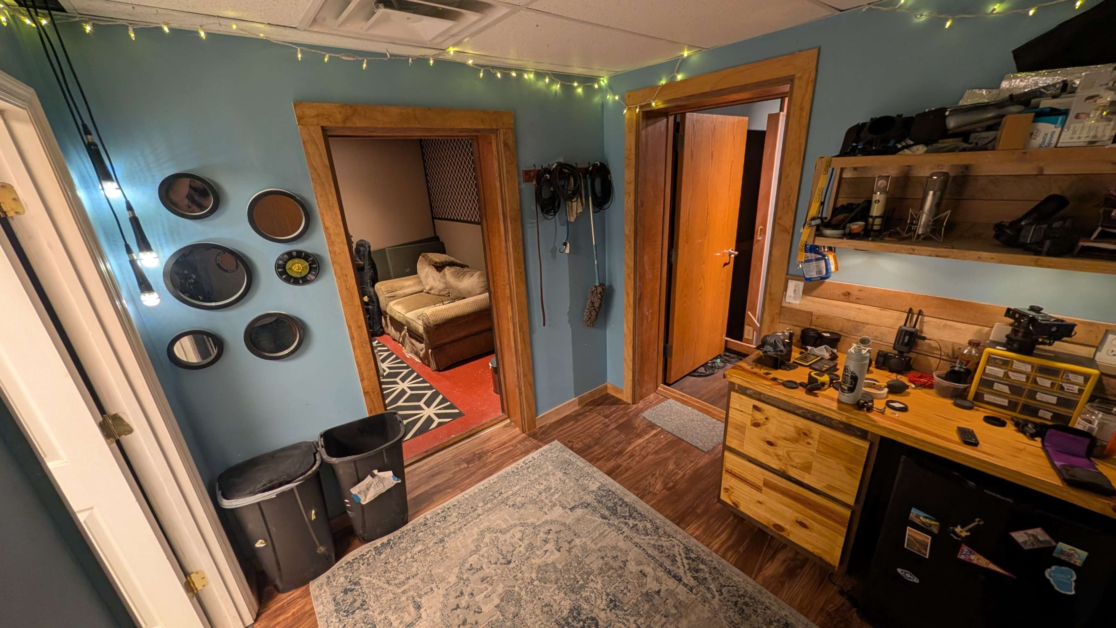 The image shows a room with blue walls, a wooden desk filled with tools and equipment, a rug on the floor, and two doorways leading to adjacent spaces.