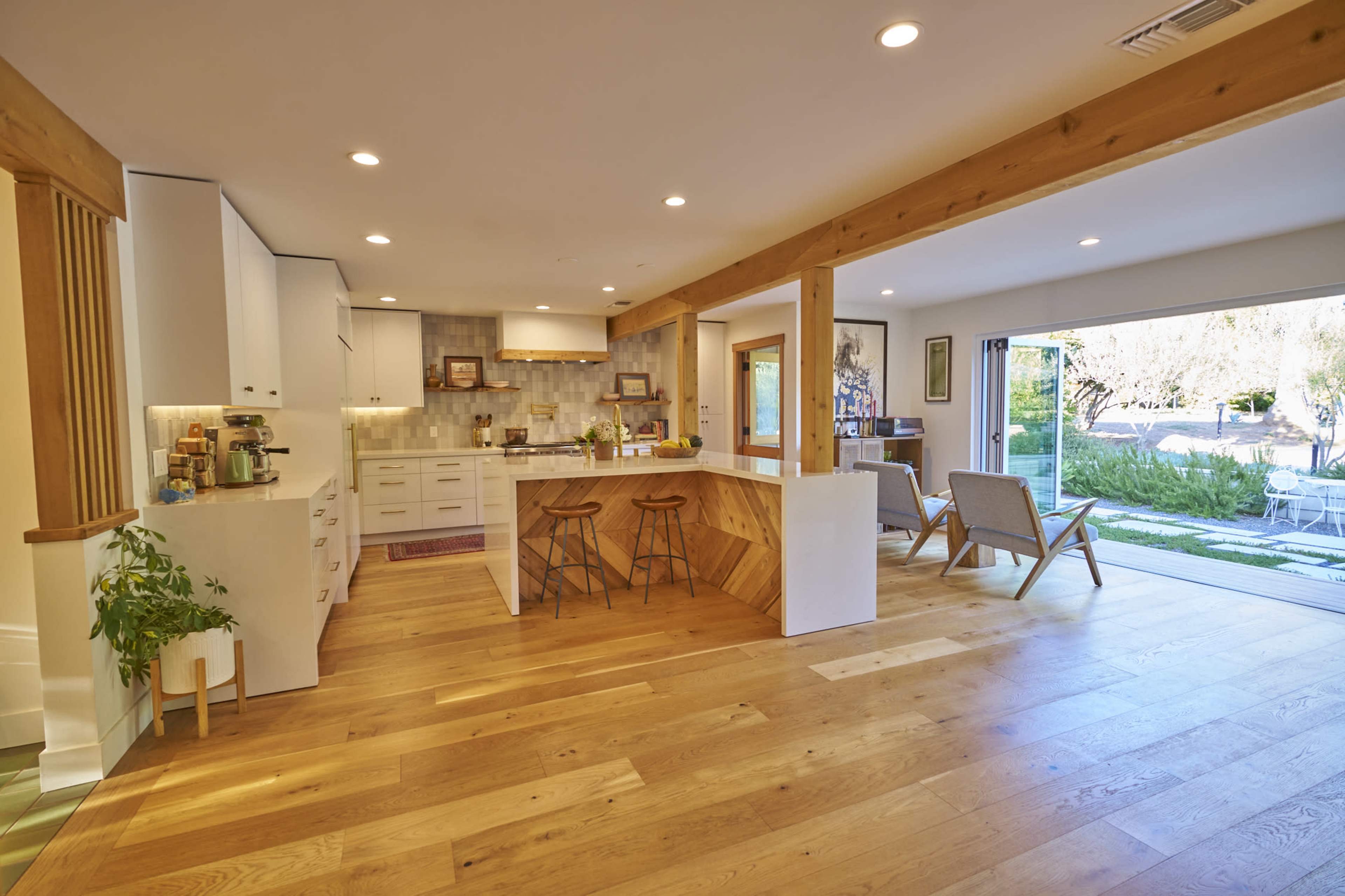 A modern kitchen features a central island with bar stools, wooden beams, and large sliding glass doors that open to an outdoor patio.