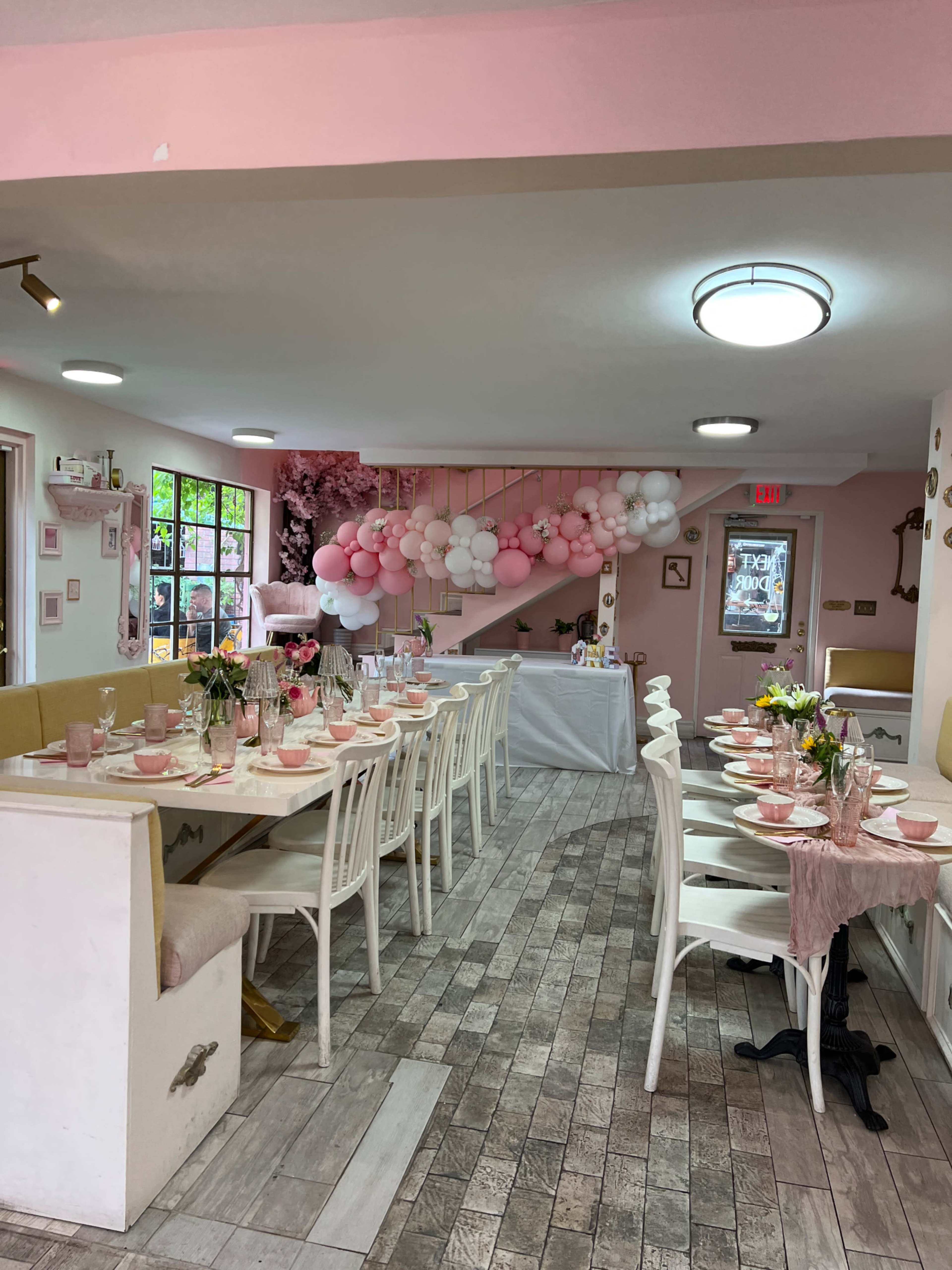 The image shows a pastel-themed dining area with white chairs and tables adorned with pink table settings and a balloon arch.