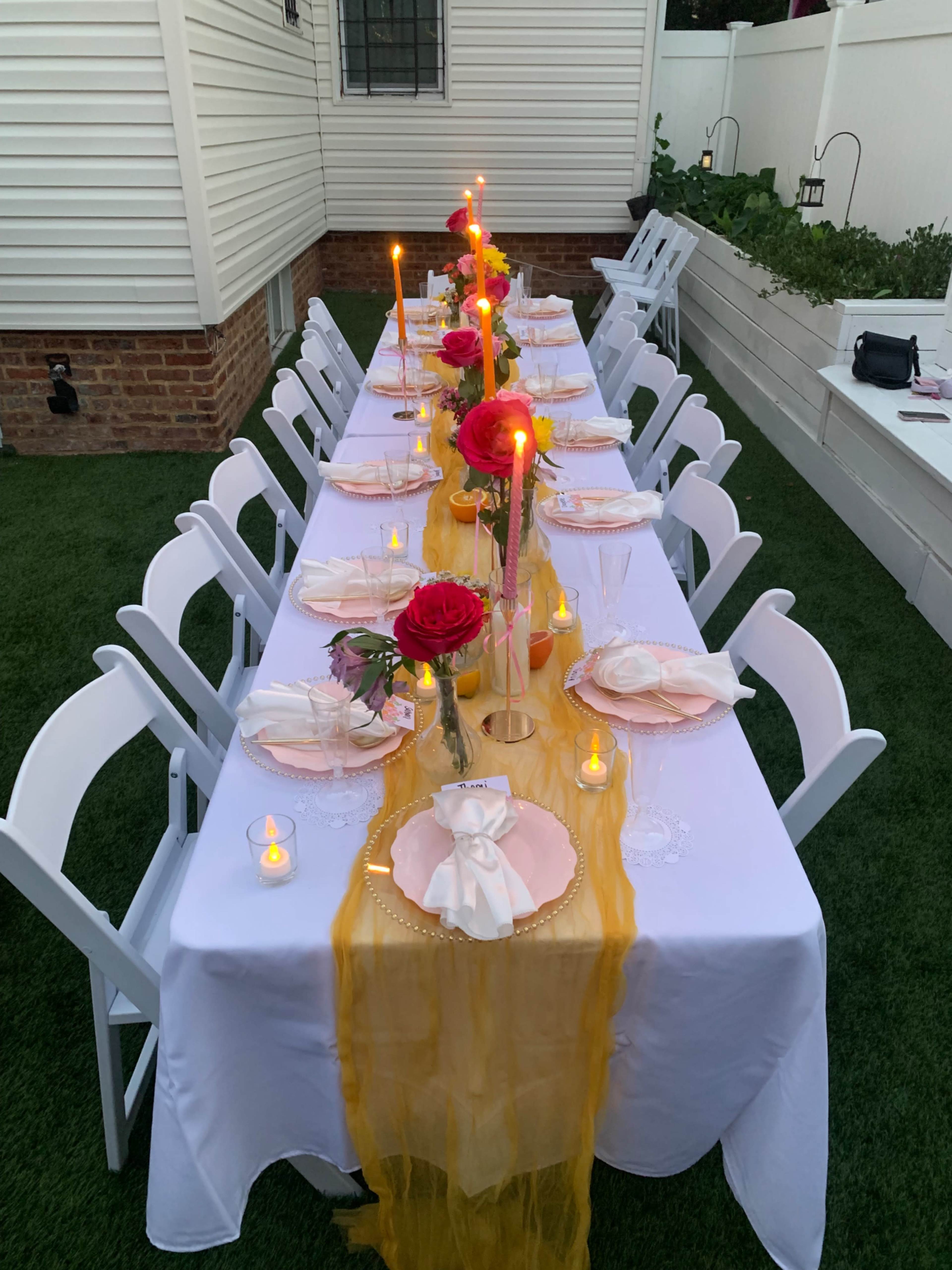 A long table is set outdoors with white chairs, decorated with yellow fabric, candles, and red roses.
