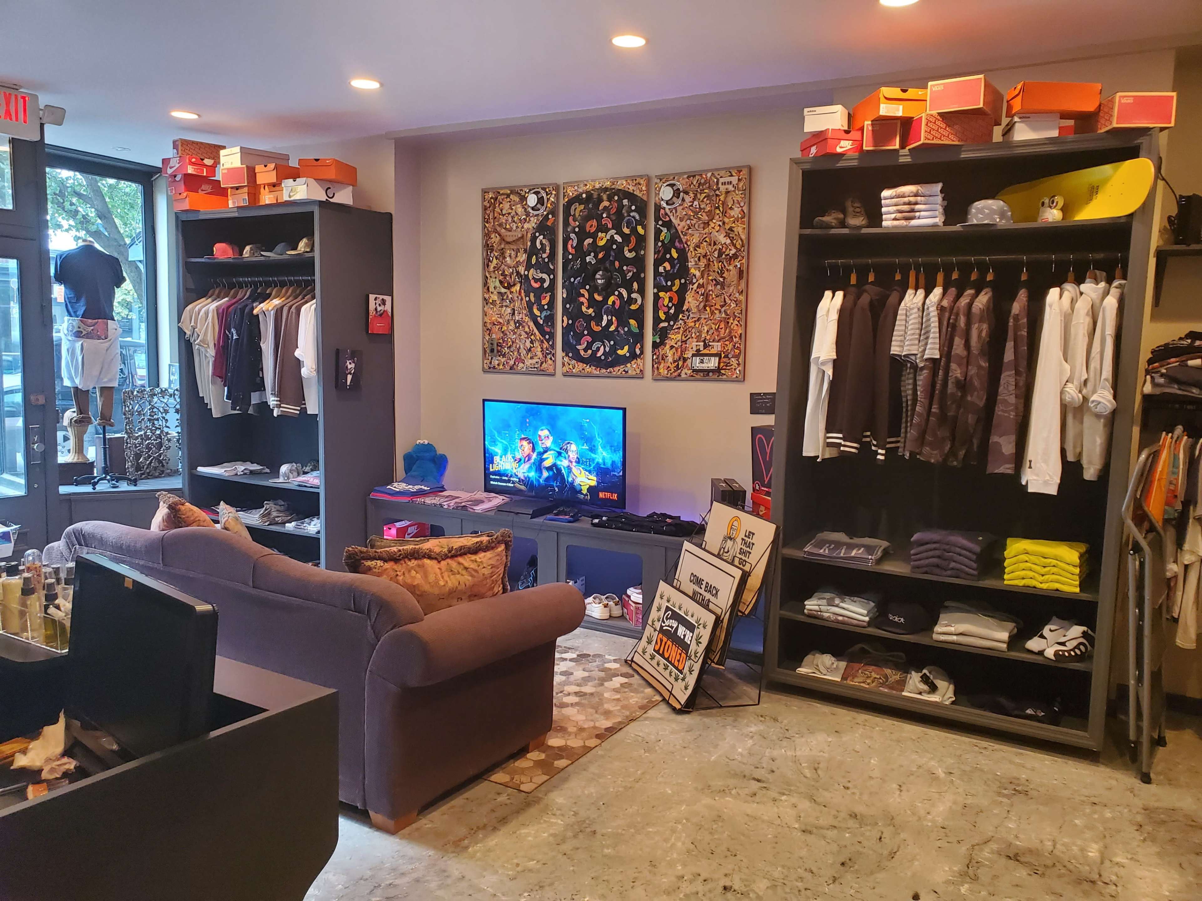 A clothing store interior featuring a sofa, racks of apparel, a television displaying colorful visuals, and various shoe boxes displayed on shelves.