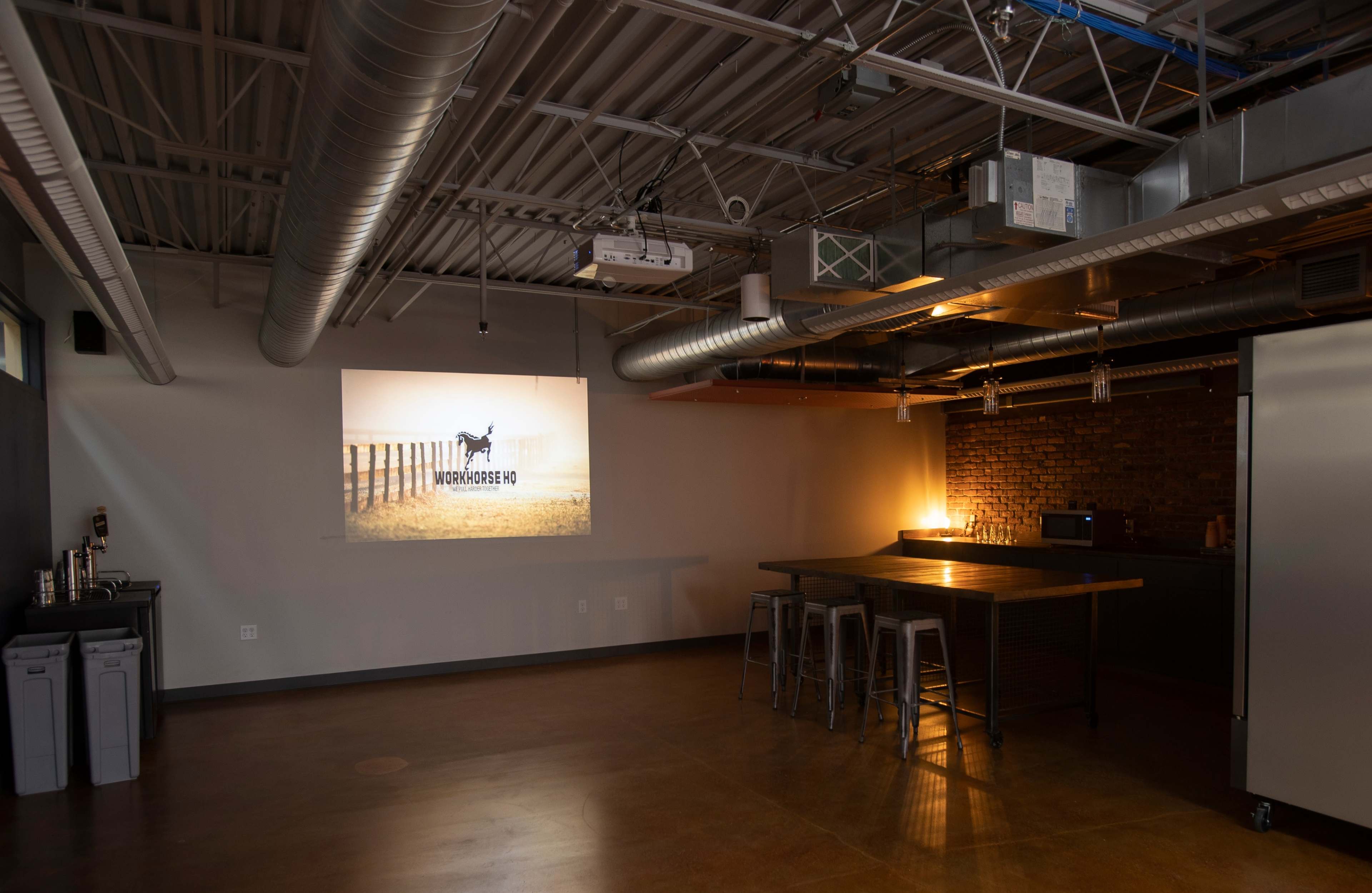 The image depicts a modern, industrial-style meeting room with a projected presentation on one wall, a kitchen area, and a central wooden table surrounded by metal stools.