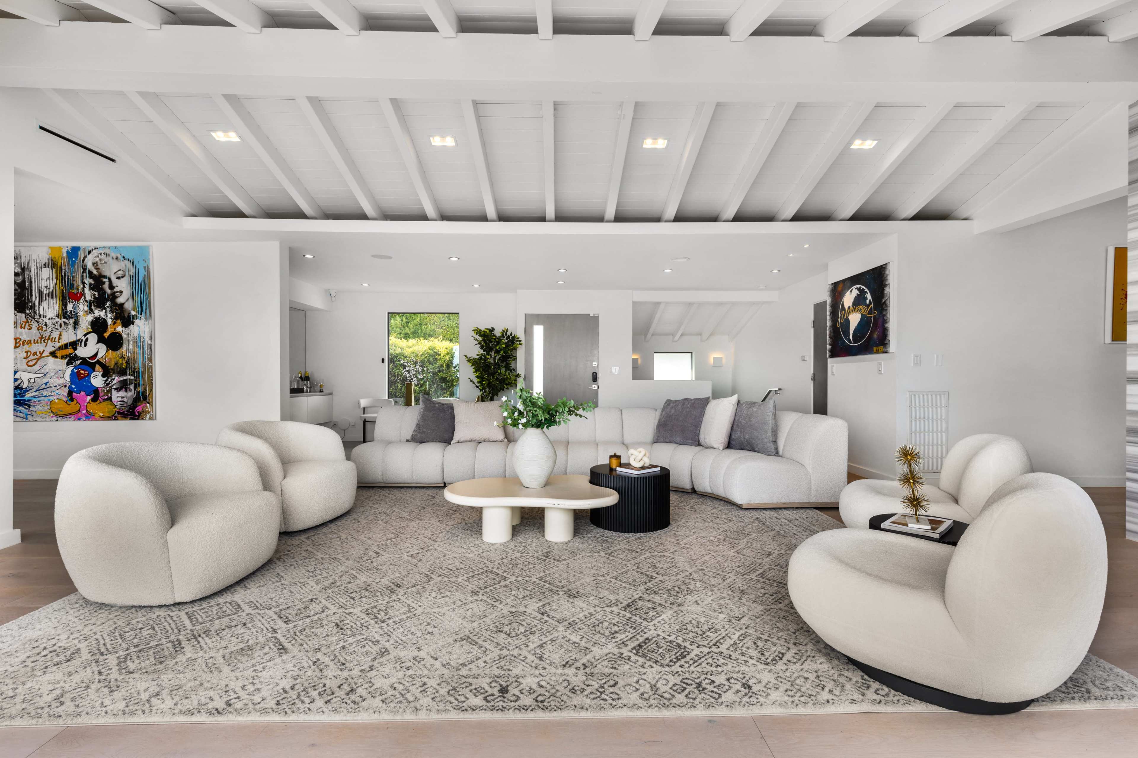 The scene depicts a modern living room featuring a large sectional sofa, two upholstered armchairs, a circular coffee table, and a patterned area rug under a beamed ceiling.