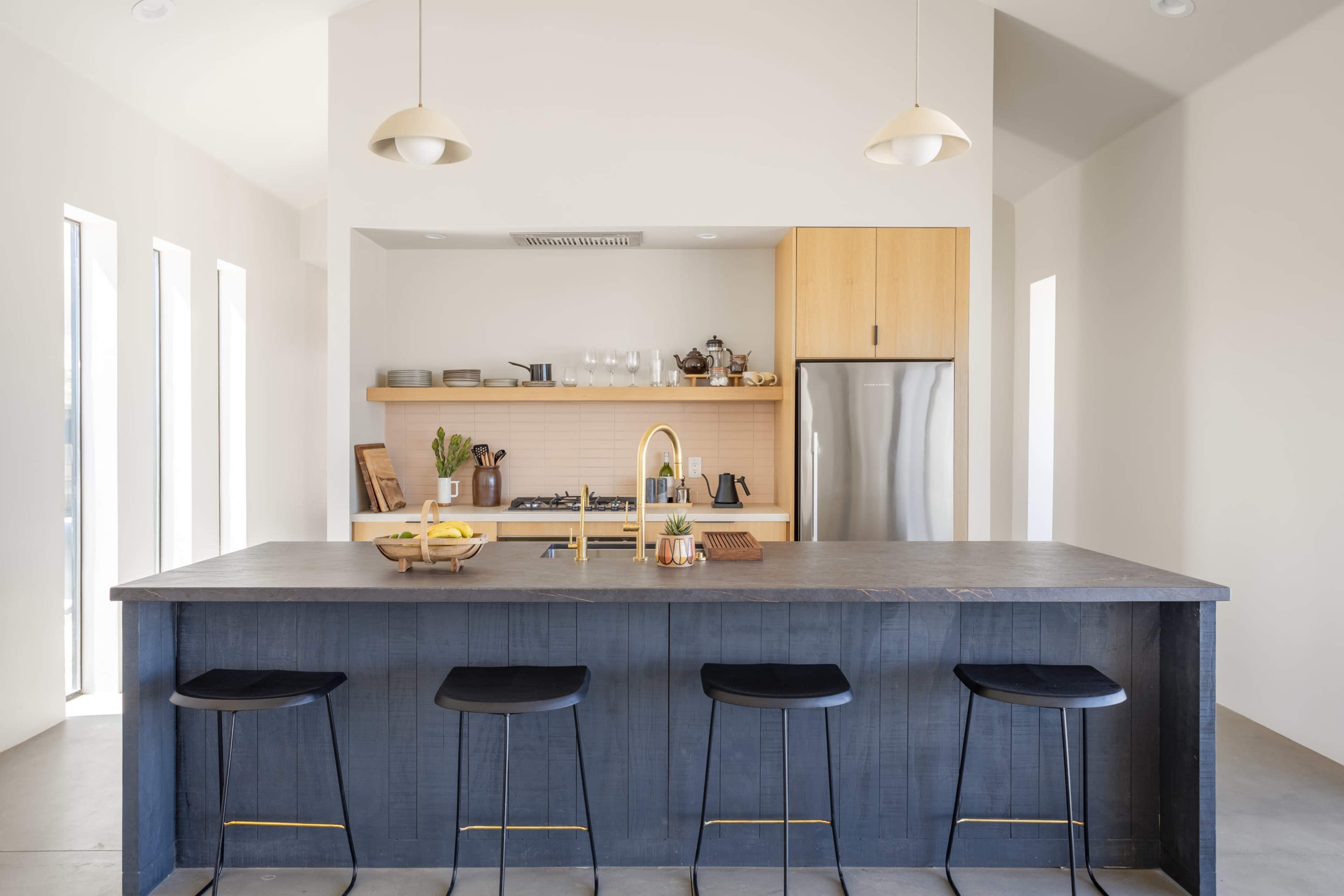 A modern kitchen featuring a large dark island with four bar stools, a stainless steel refrigerator, and light wood cabinetry.