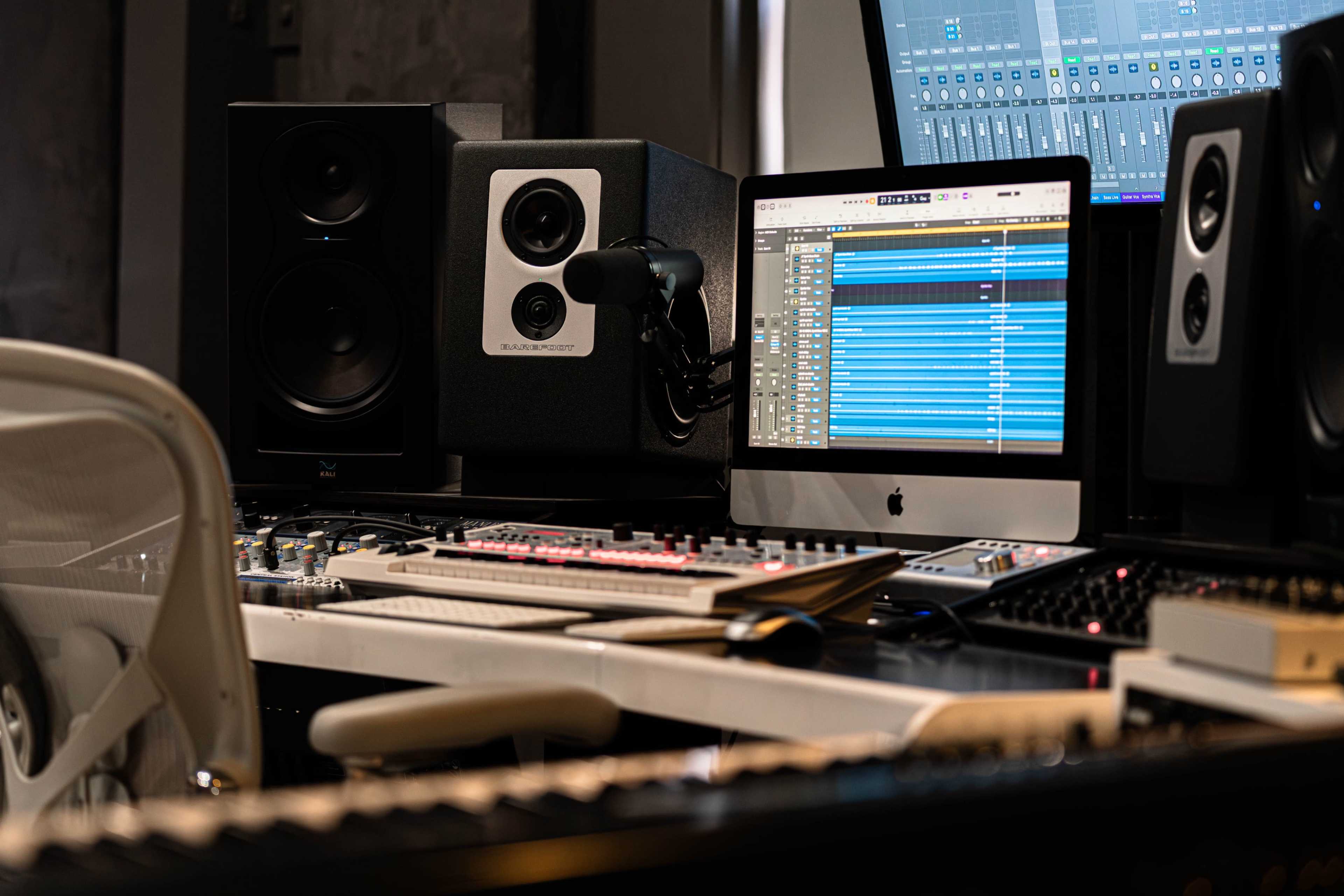 A music production workspace features a computer monitor displaying audio tracks, studio monitors on either side, and a keyboard in the foreground.