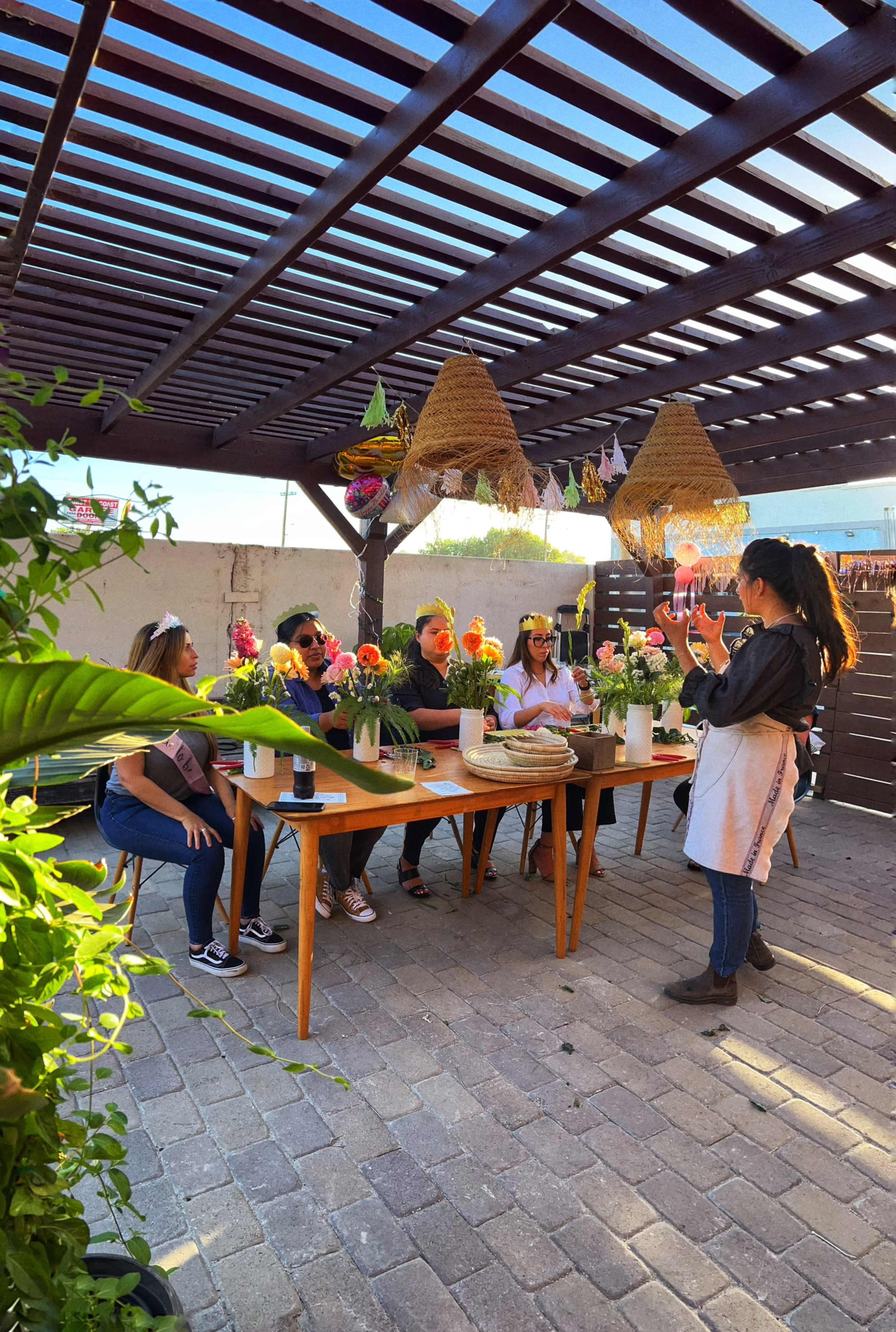 A group of five people sits at a wooden table under a covered patio while a woman stands and gestures, presumably leading a floral arrangement workshop.