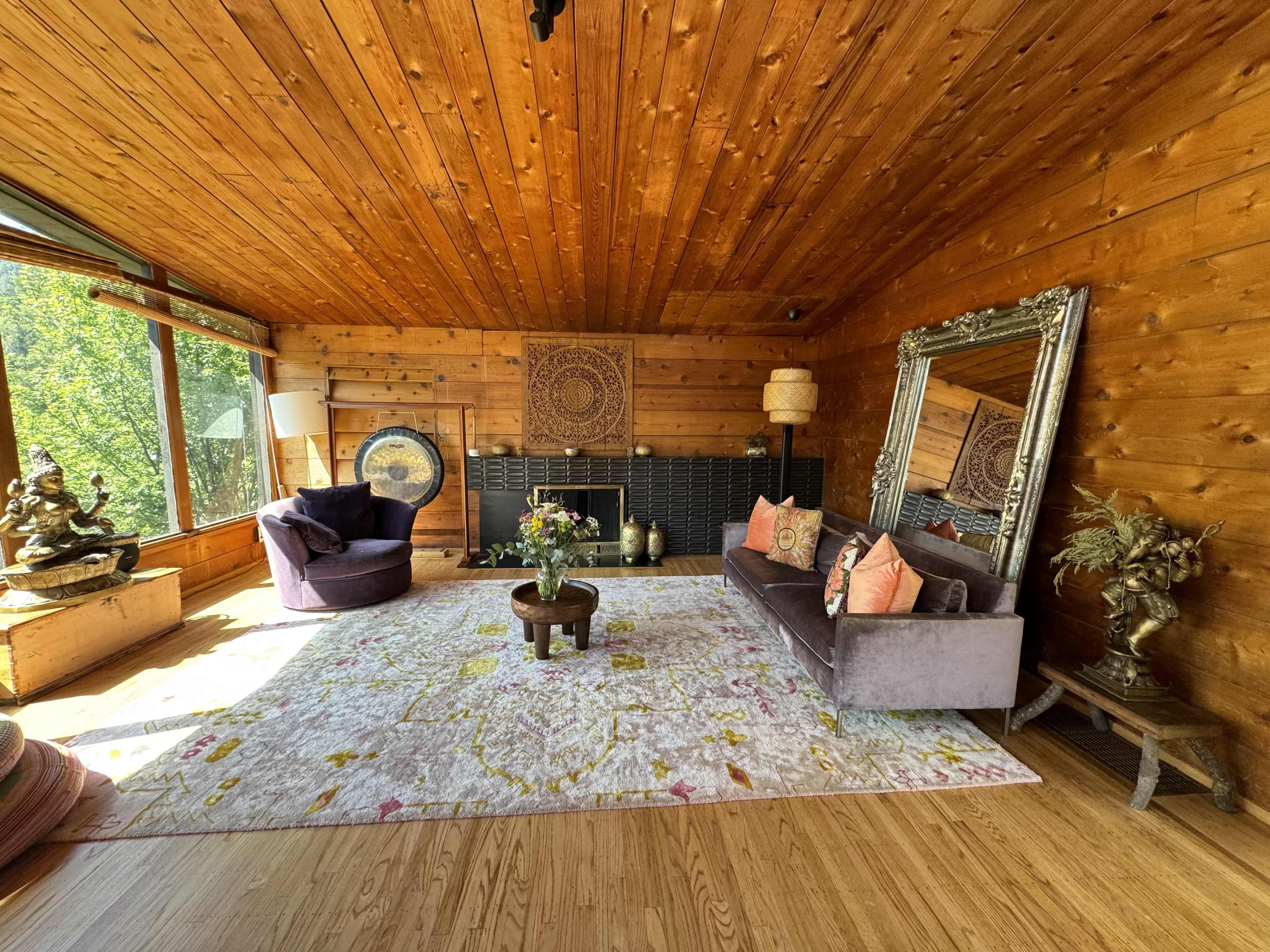 A wooden-paneled living room with a purple sofa, a chair, a large mirror, a decorative rug, and large windows allowing natural light.