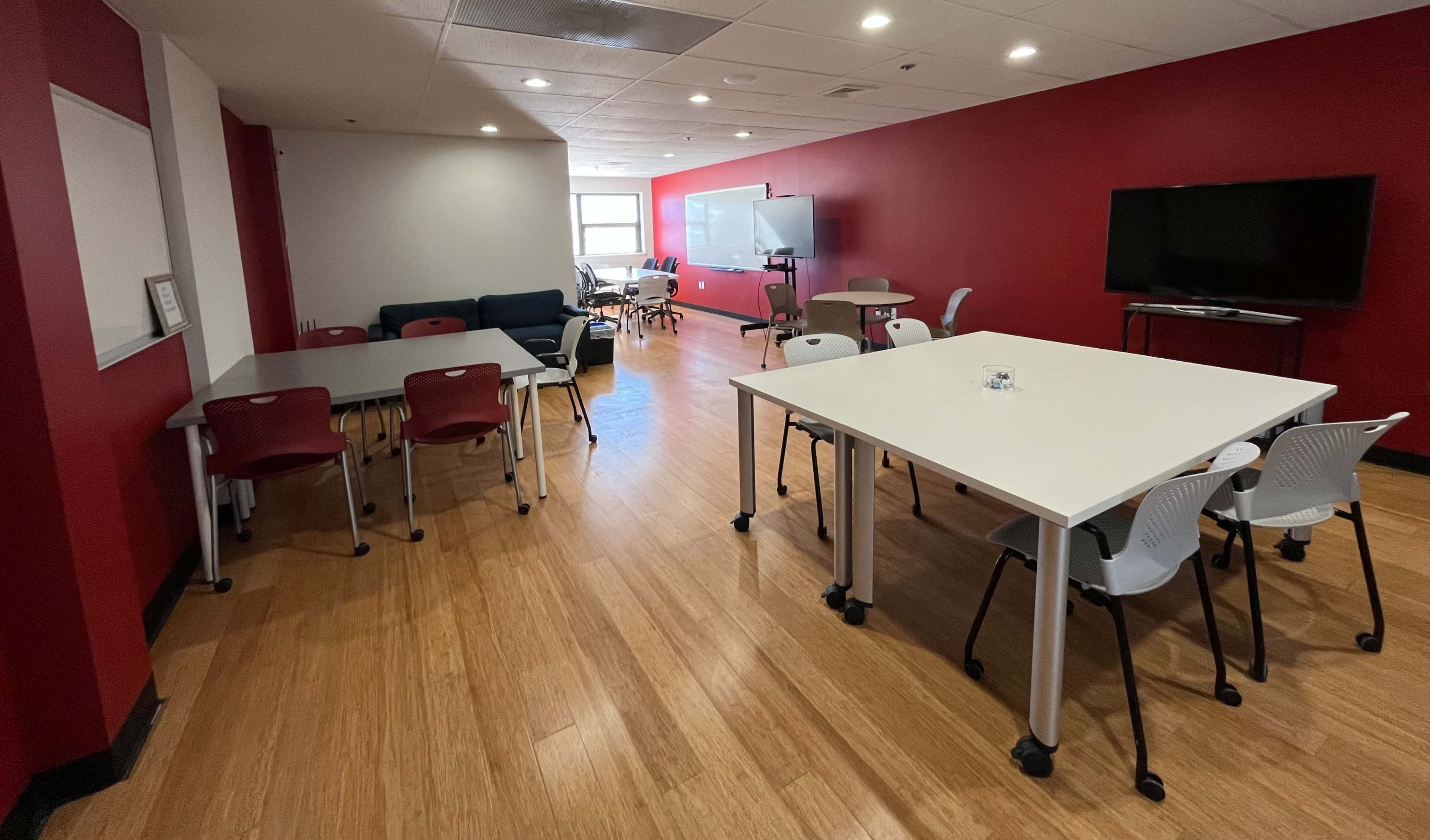 The image shows a modern, well-lit collaborative workspace featuring several tables and chairs, with a red accent wall and a gray couch in the background.