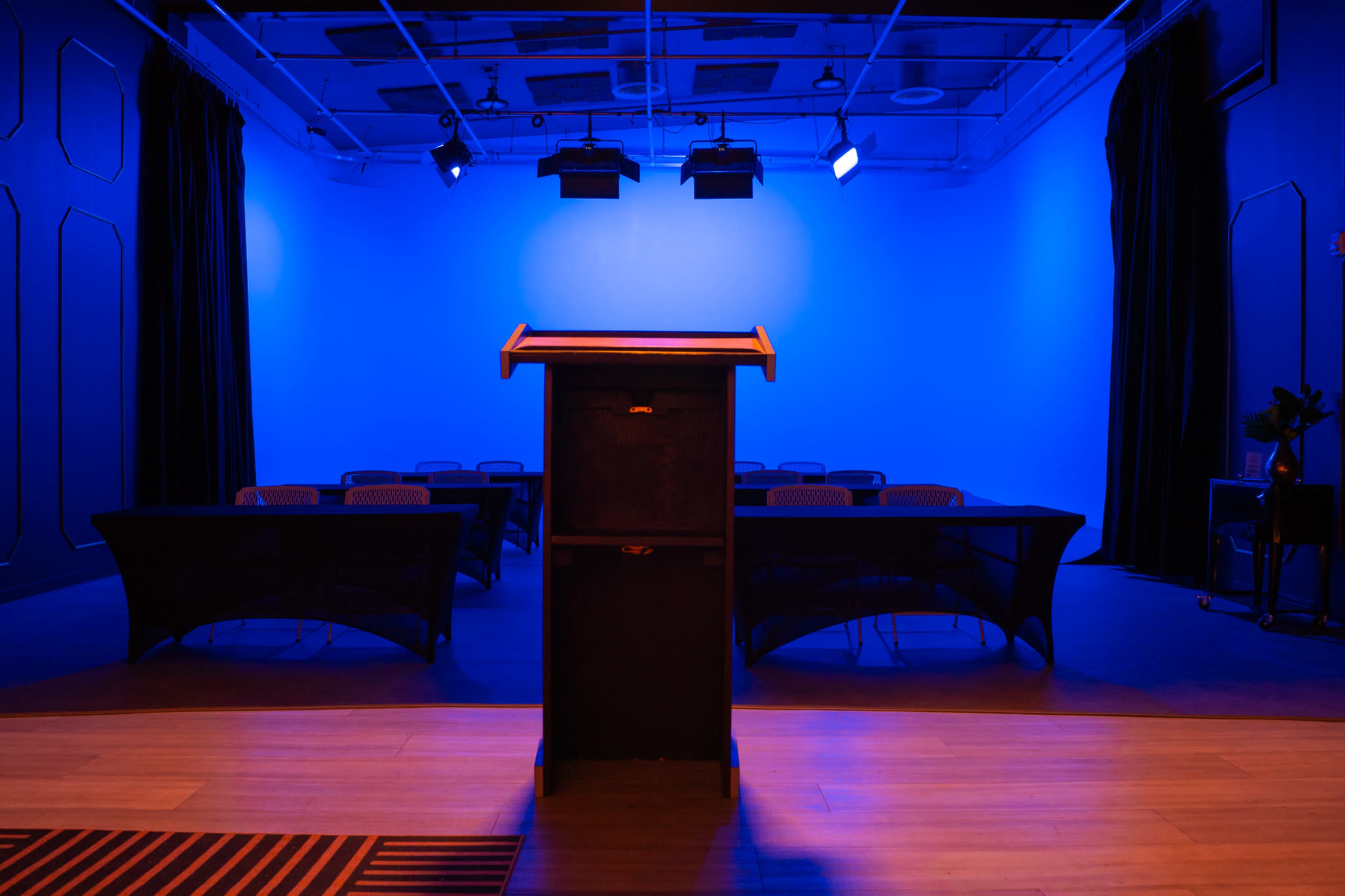 A podium is positioned at the forefront of a dimly lit room, featuring blue lighting and several empty tables arranged for an event.
