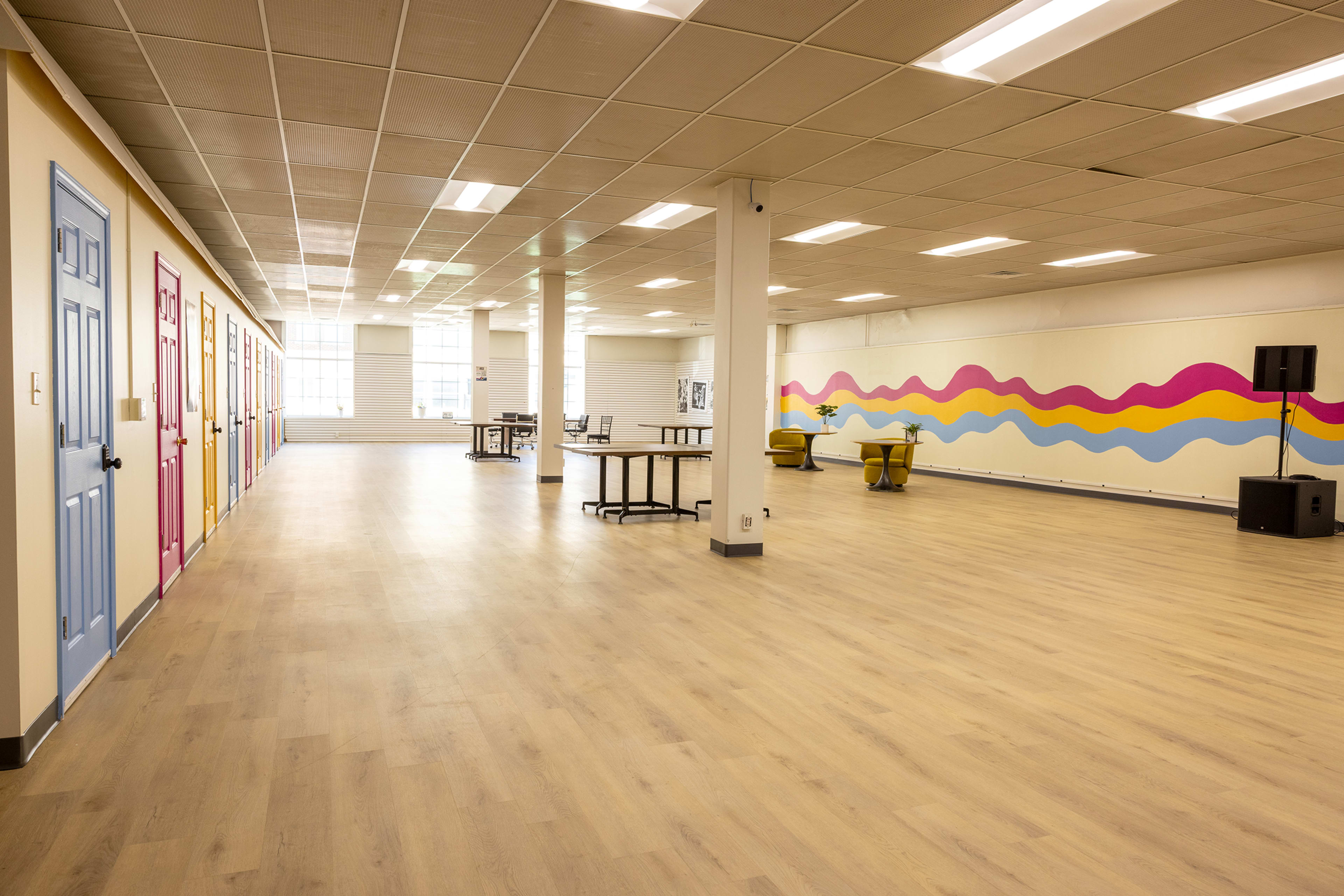 The image shows a spacious, brightly lit room with colorful doors lining one wall and a vibrant wave mural on the opposite wall.
