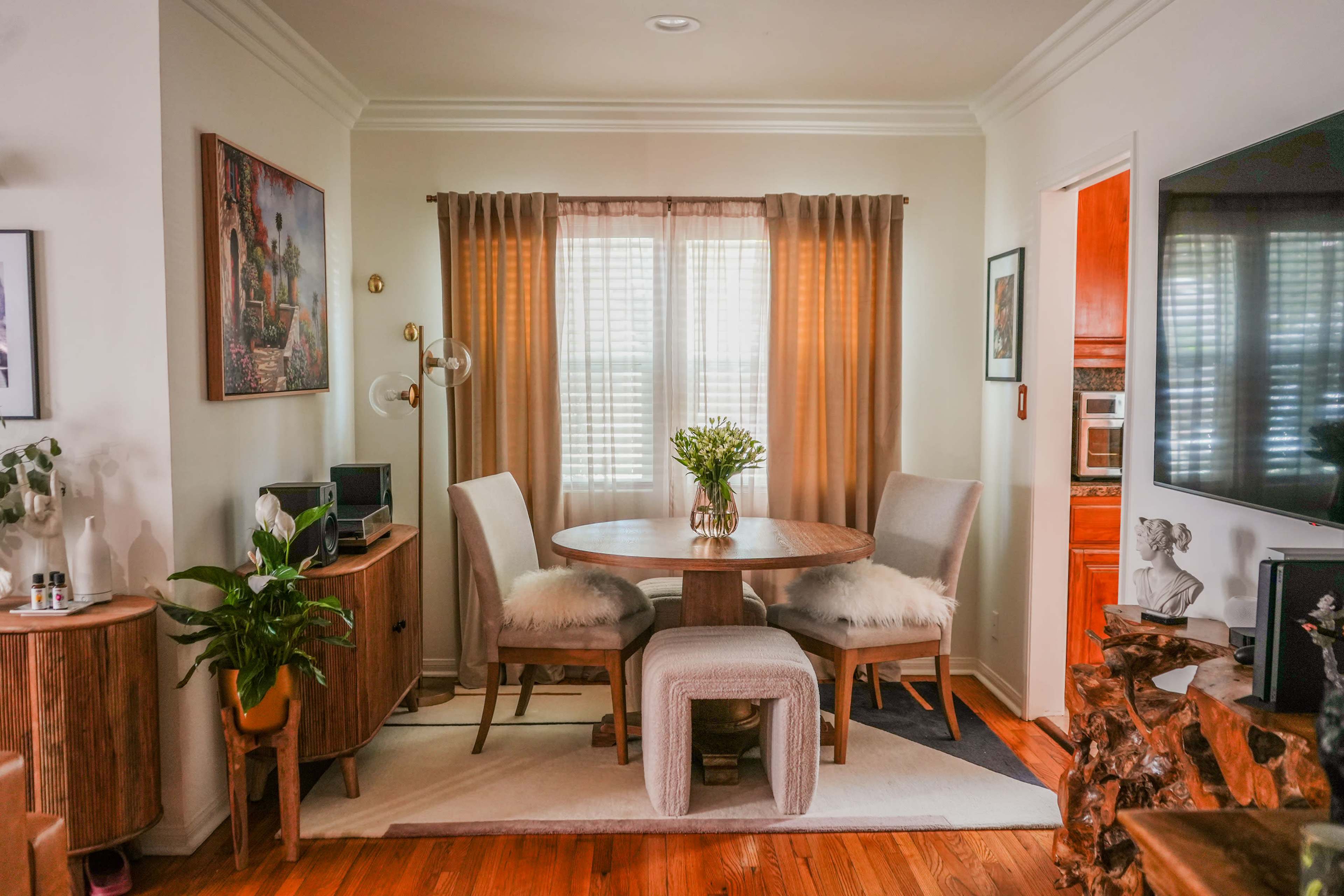 A small dining area features a round wooden table with two upholstered chairs, a potted plant, and sheer curtains allowing light to filter in.