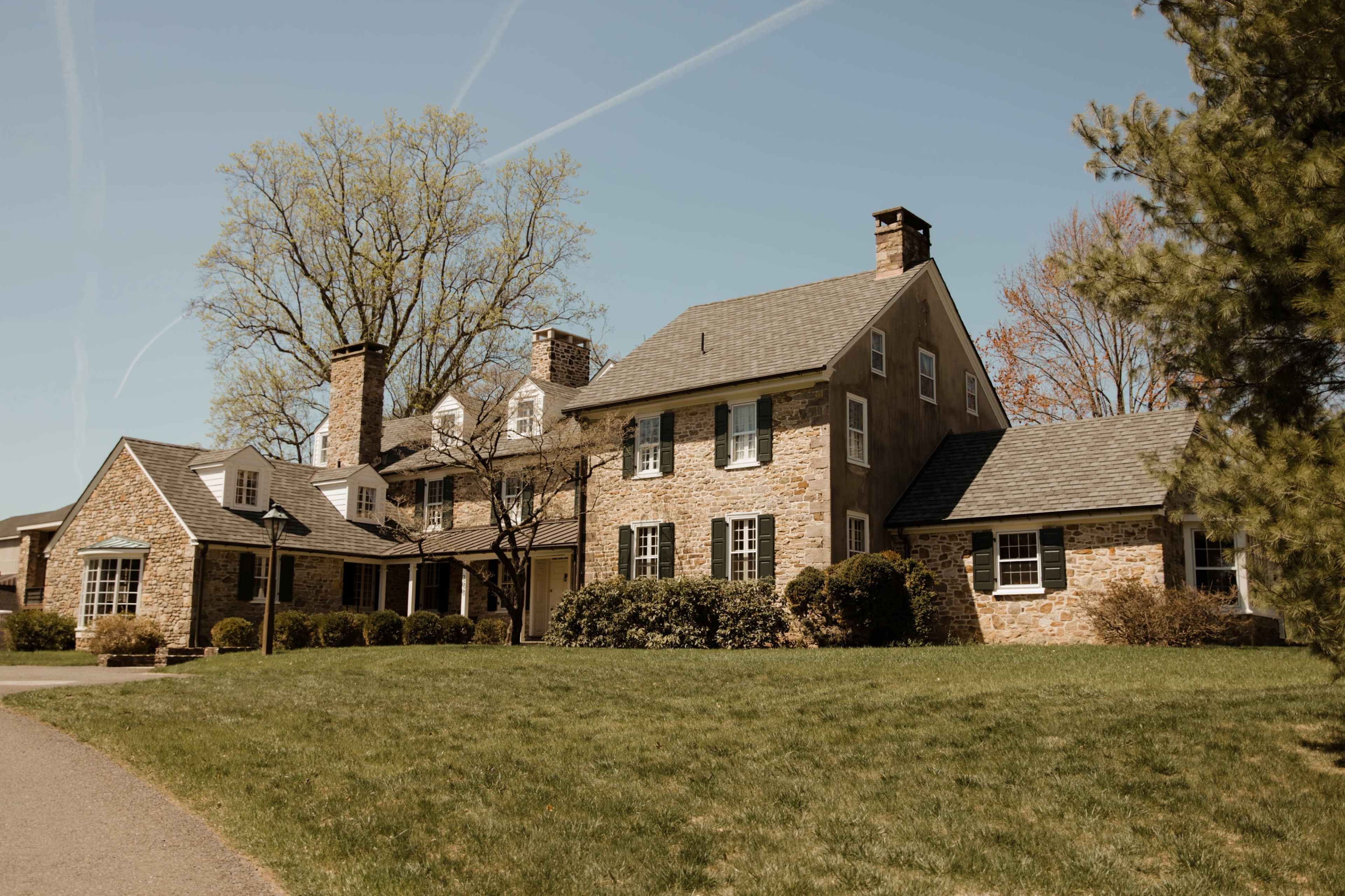The image shows a large, two-story stone house with multiple chimneys, surrounded by a green lawn and trees.