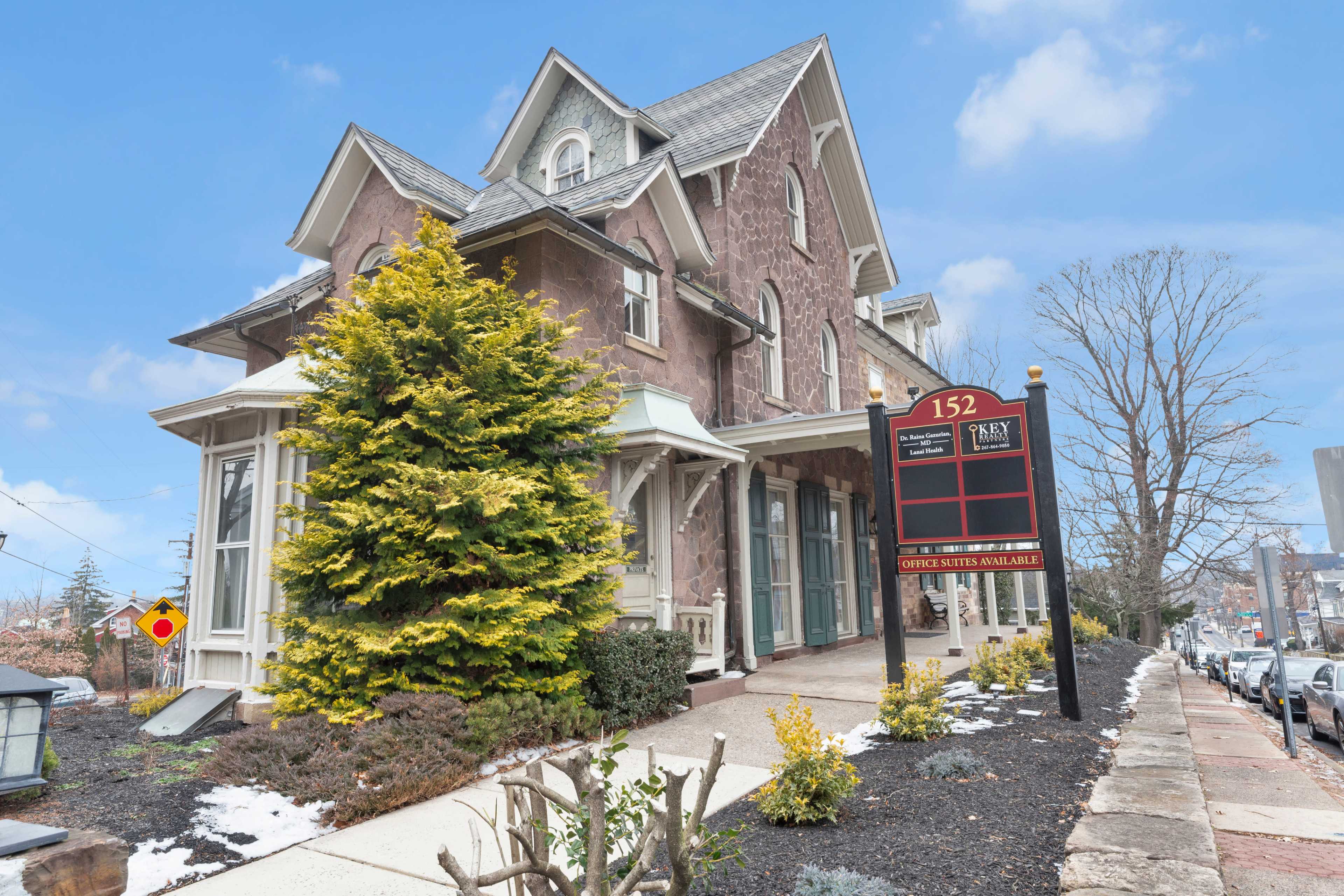 A large, stone building with multiple gables features a well-manicured front yard and a prominent sign displaying available rental information.