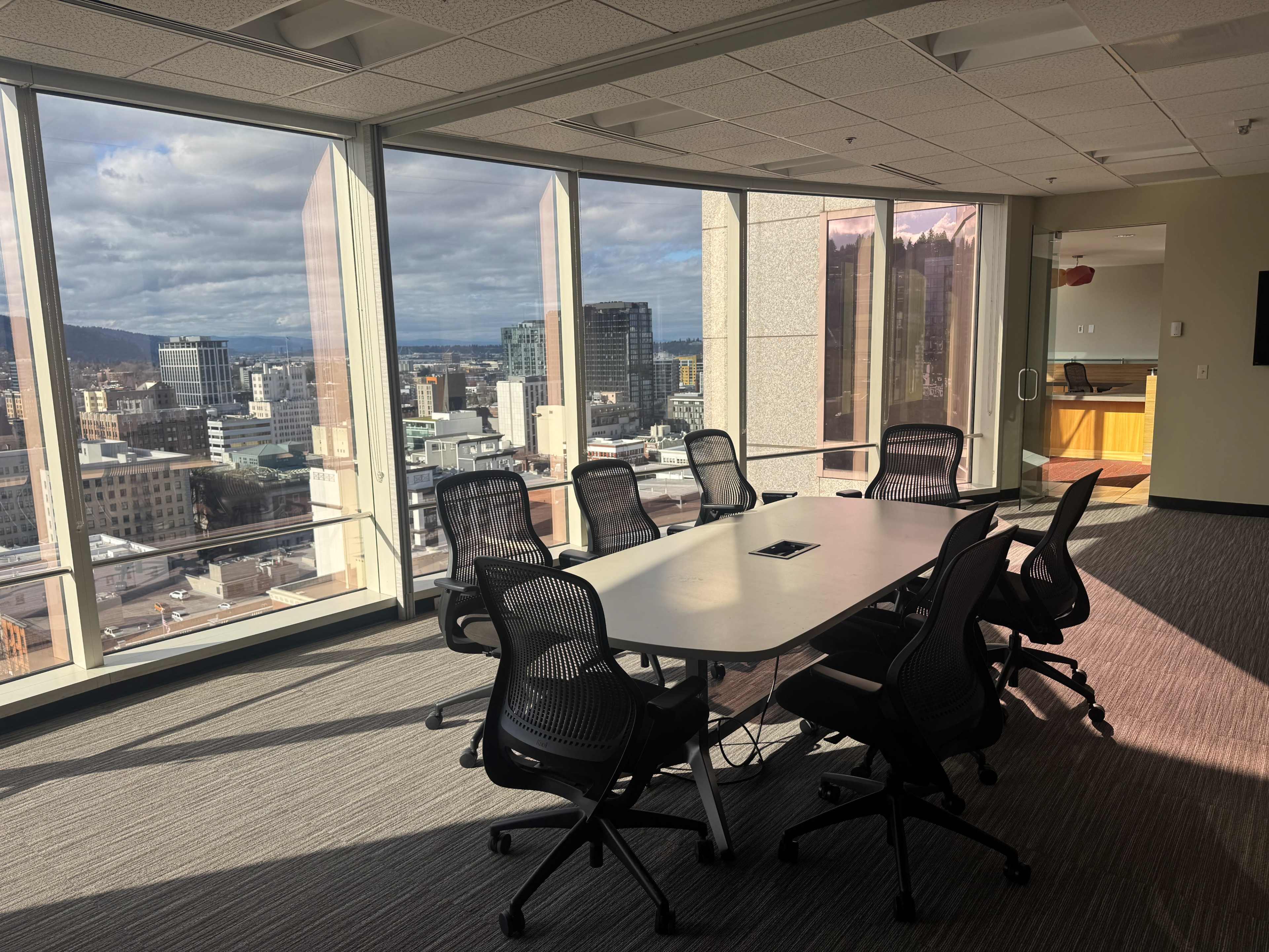 A modern conference room features a rectangular table surrounded by chairs, with large windows showcasing a cityscape.