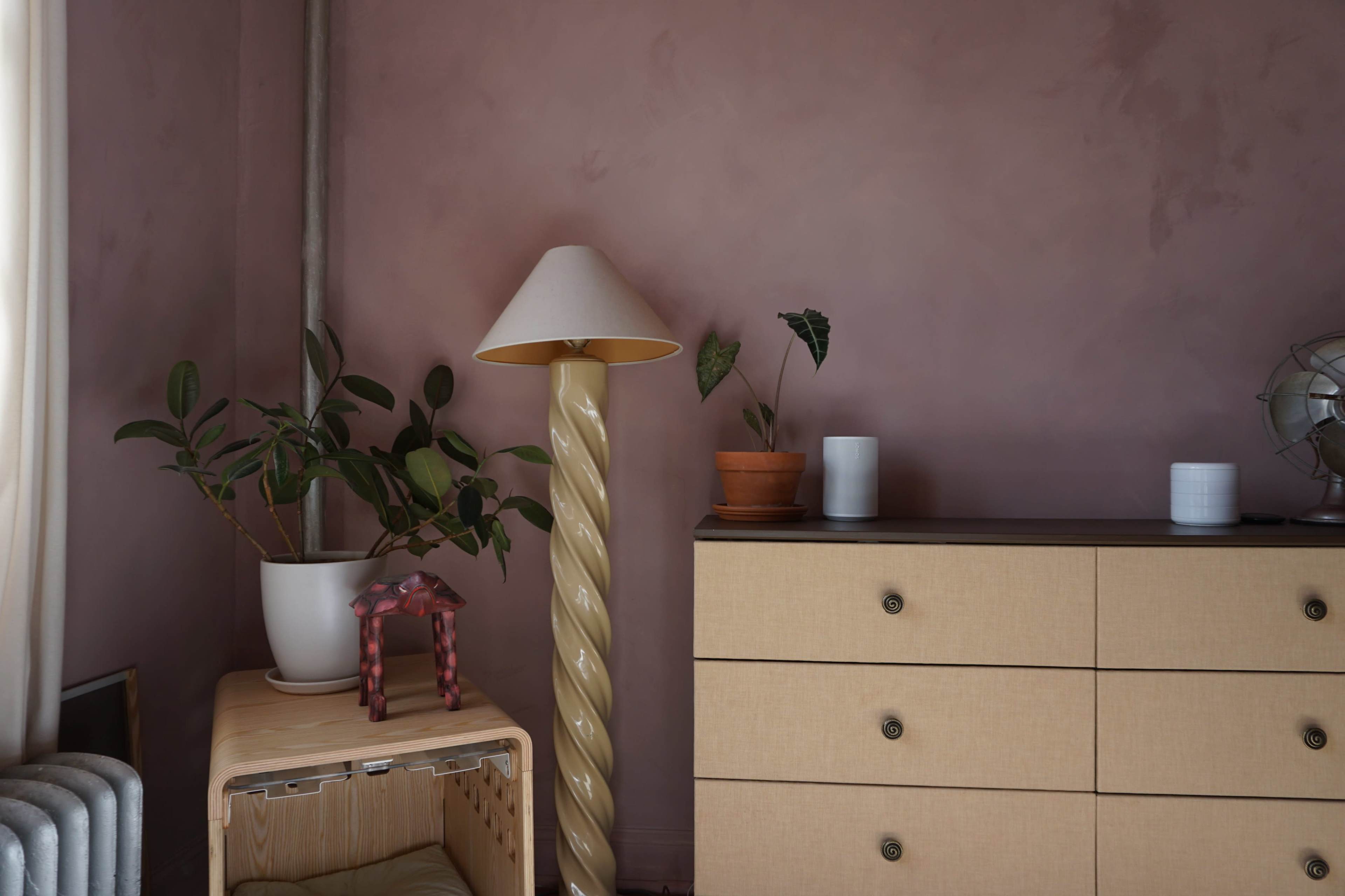 A cozy interior scene features a beige dresser, a spiral lamp, potted plants, and decorative items against a softly colored wall.