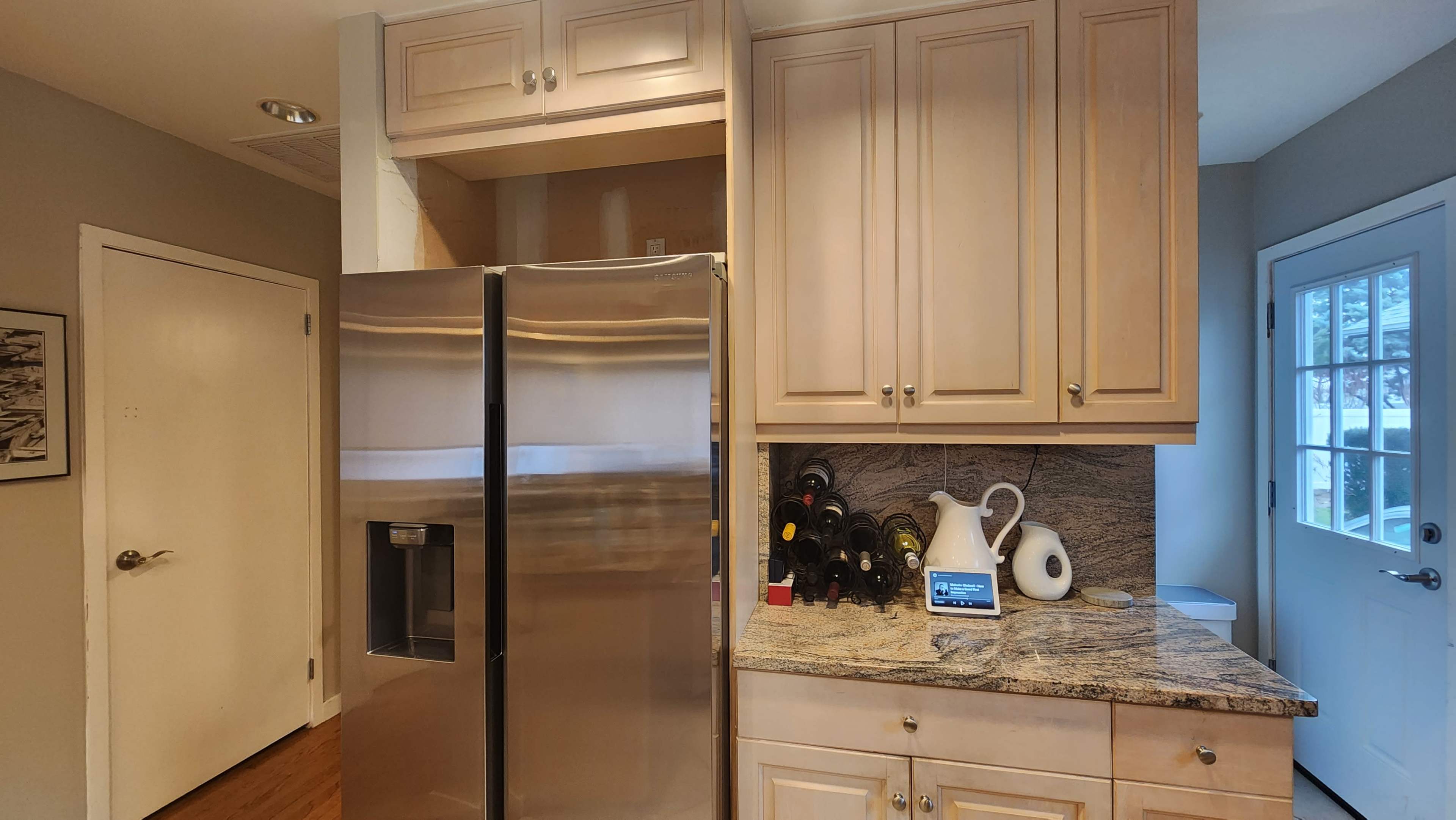 A kitchen area featuring a stainless steel refrigerator, wooden cabinets, a granite countertop with several wine bottles, and a digital display on the counter next to a decorative pitcher.