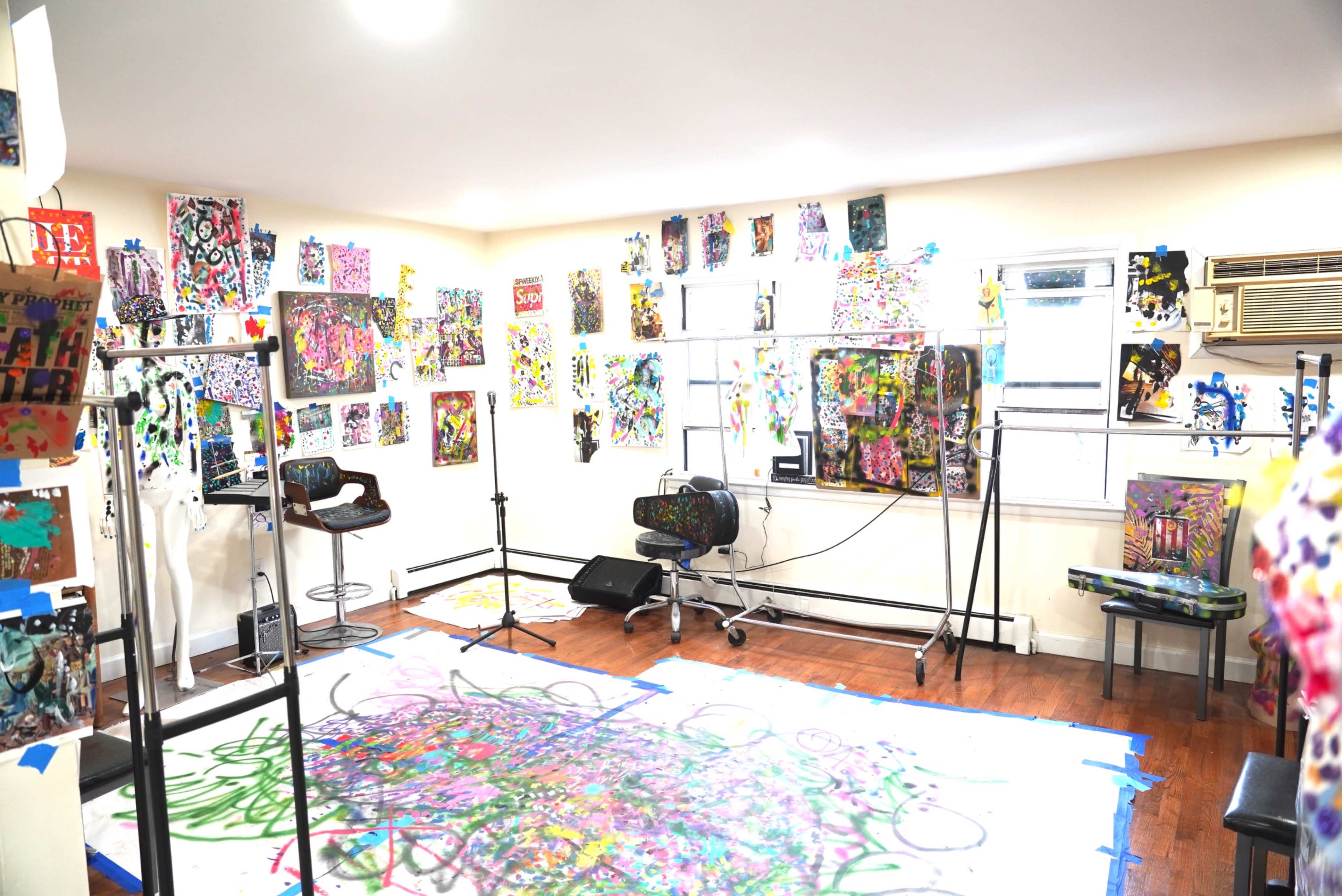 The image shows a brightly colored art studio filled with various pieces of artwork on the walls and scattered paint-covered surfaces, with easels and tools arranged throughout the space.