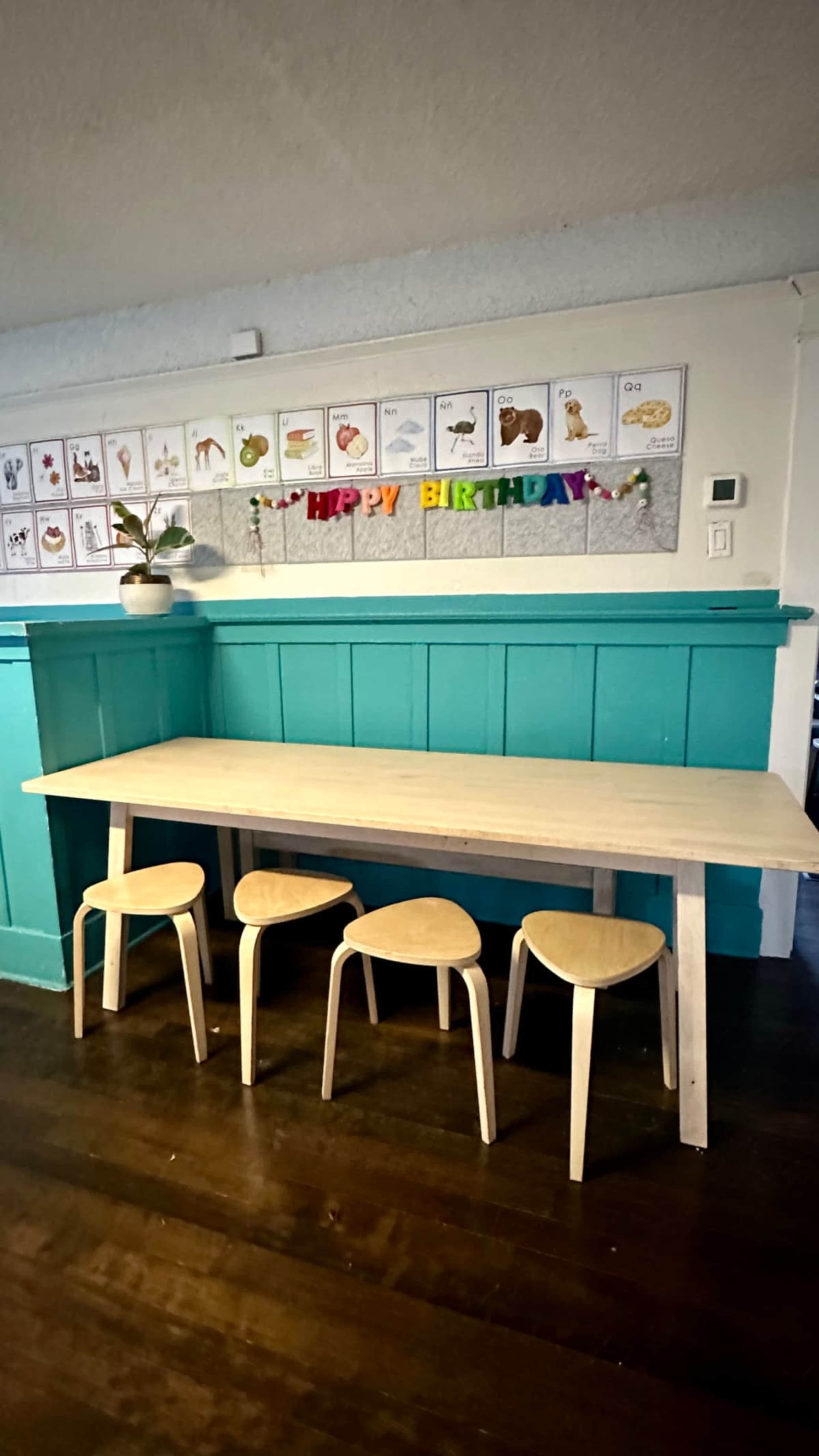 A light wooden table with four matching chairs is positioned against a teal wall, adorned with birthday decorations and educational posters.