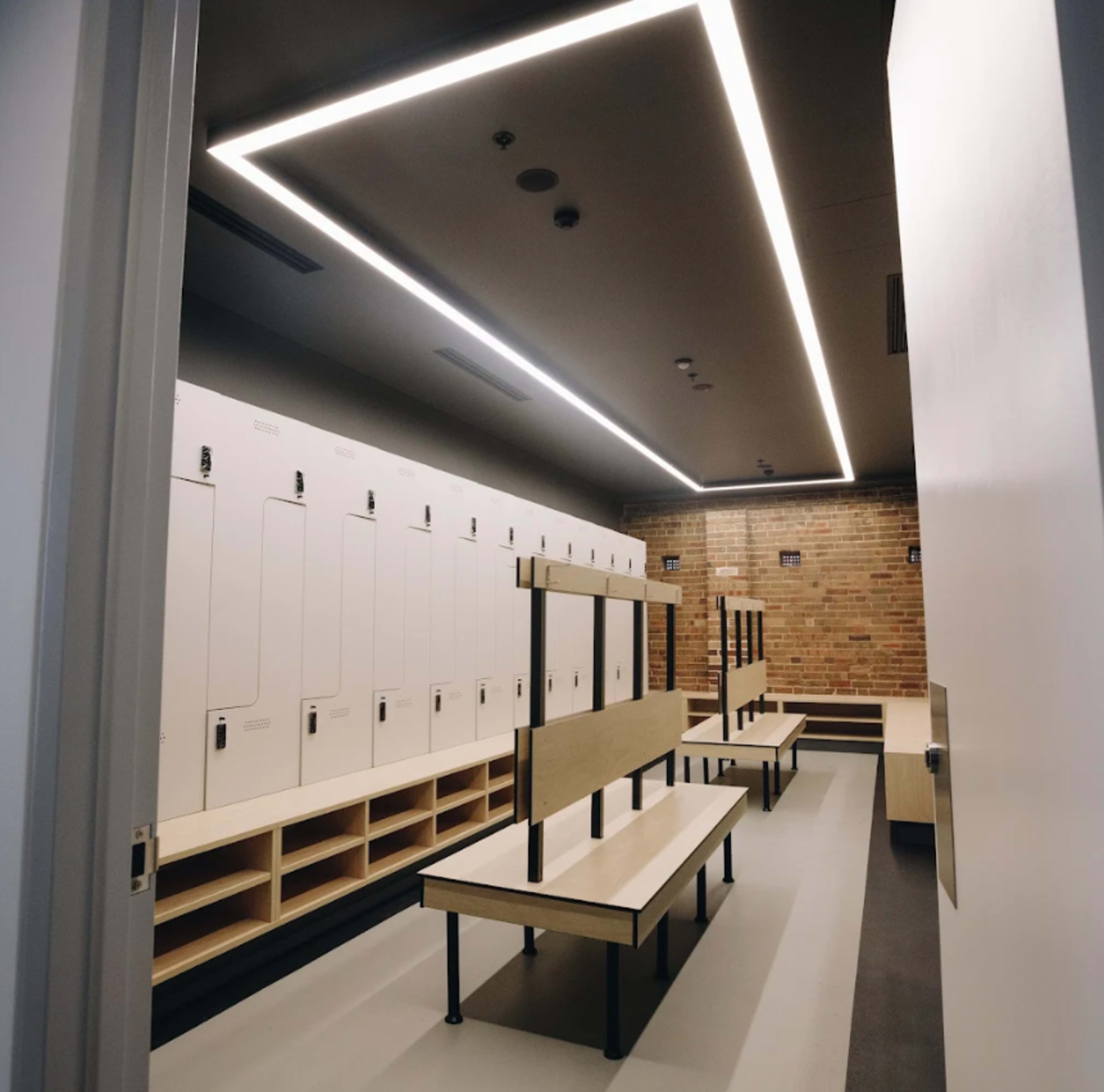 The image shows a modern locker room with wooden benches and individual lockers, illuminated by overhead lighting against a brick wall.