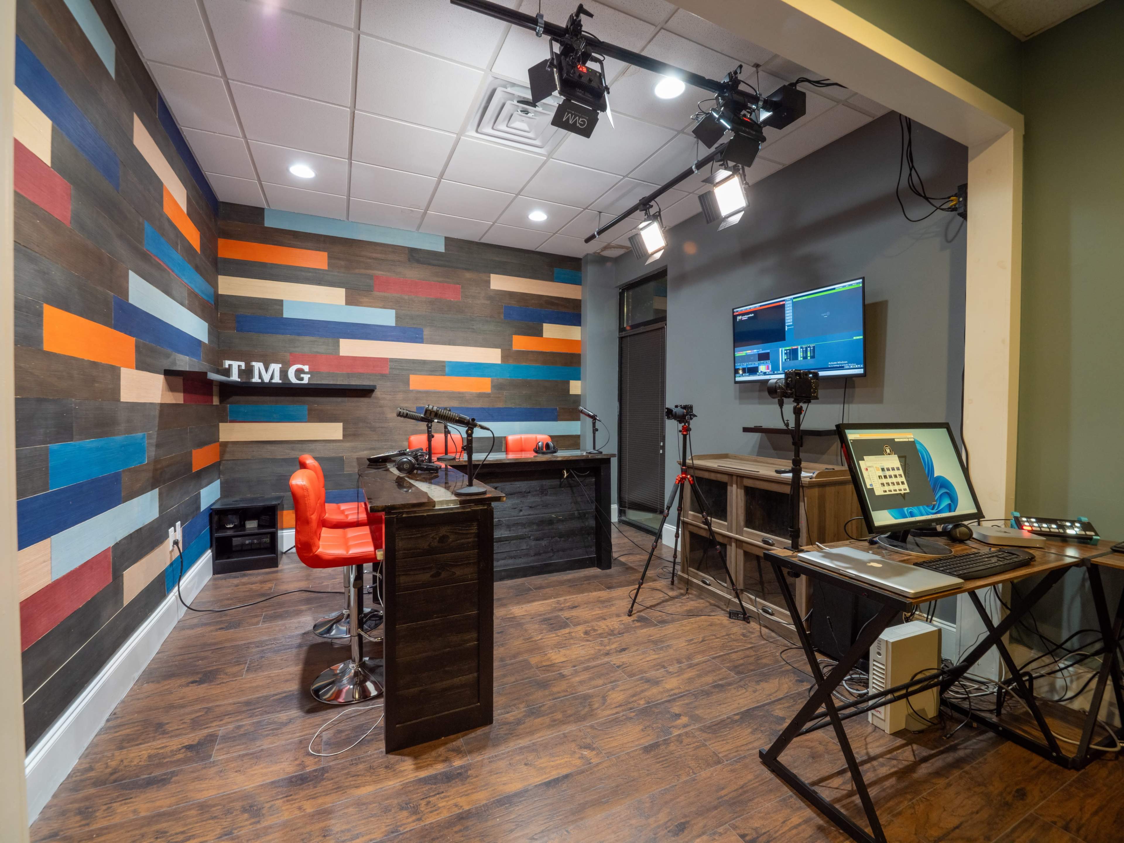 A modern recording studio with a colorful wall, a desk with two red chairs, and equipment positioned for video production.