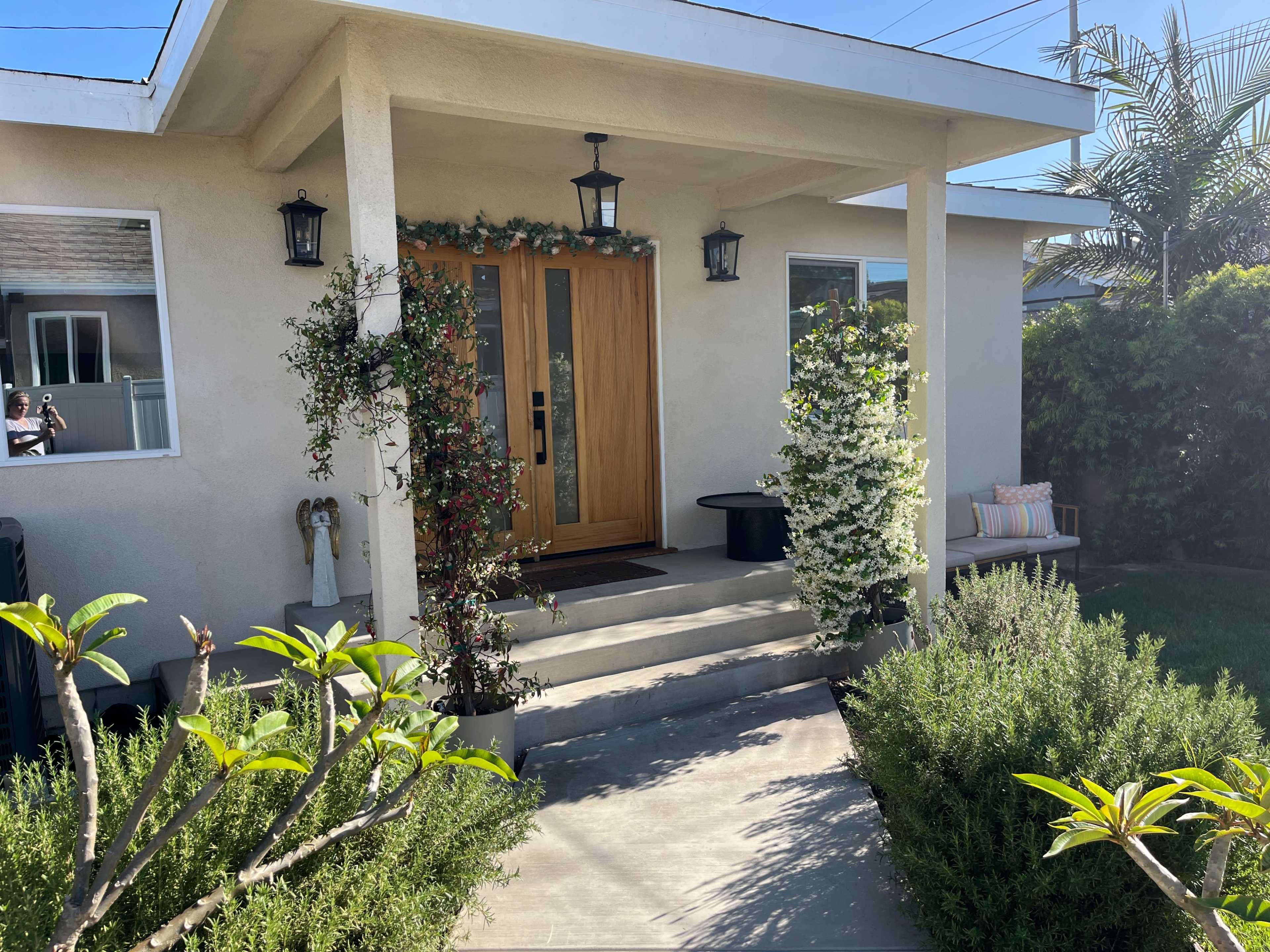 The image shows the entrance of a house with double wooden doors, flanked by potted plants and a small seating area on the porch.