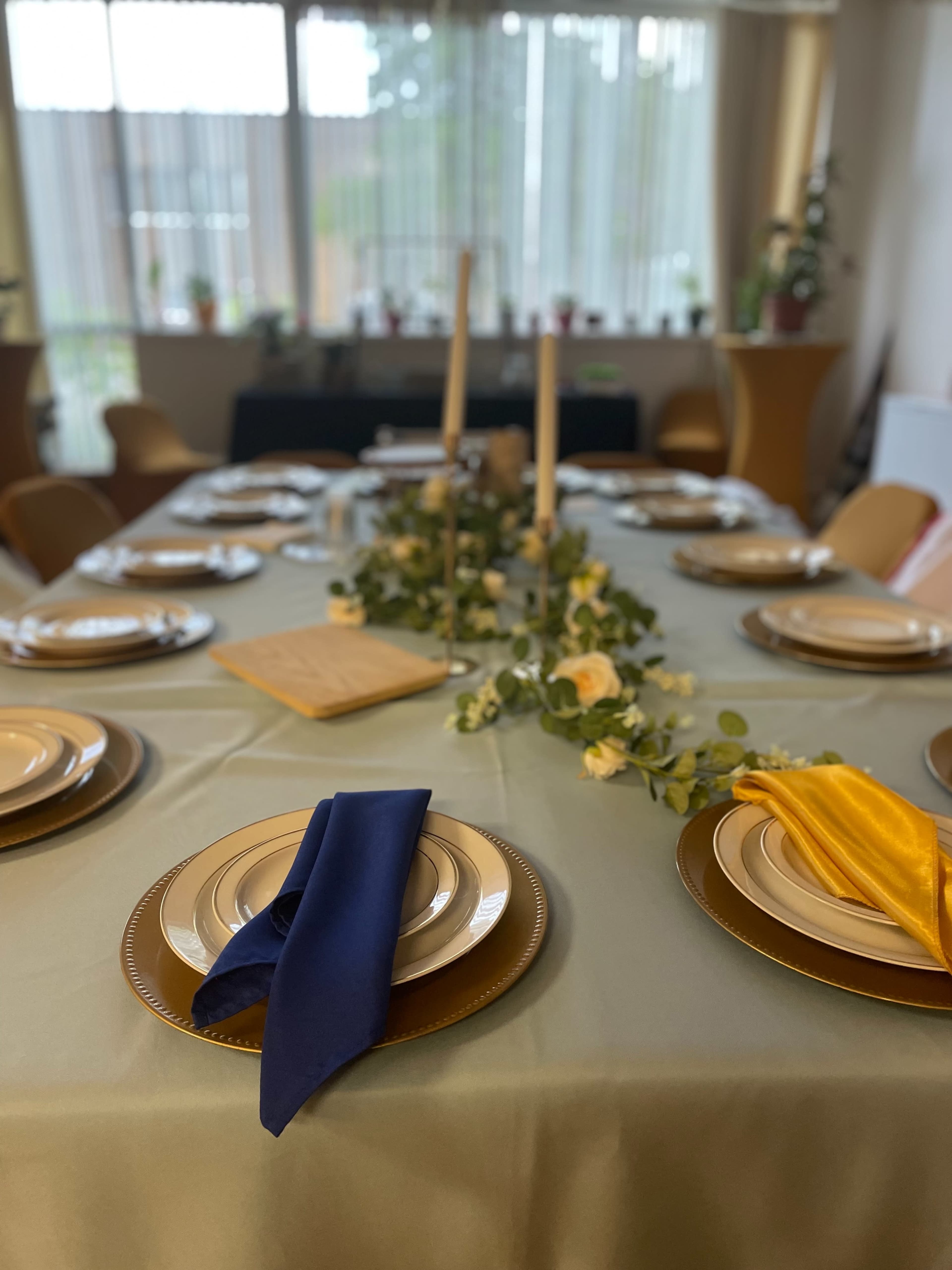 A dining table is elegantly set with plates, colored napkins, candles, and a floral centerpiece, all arranged beneath bright natural light from large windows.