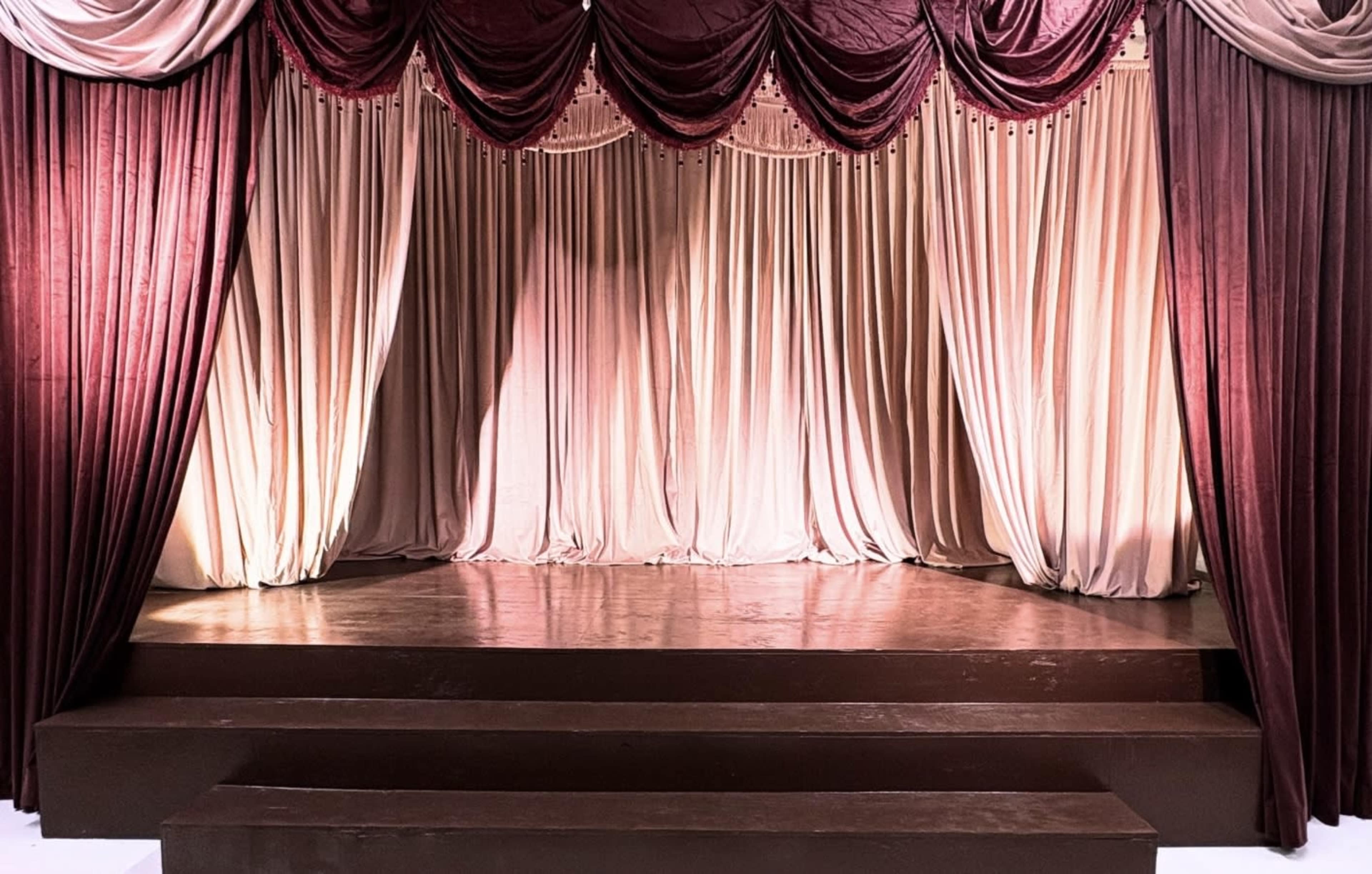 The image shows an elegantly draped theater curtain in shades of burgundy and beige, set against a wooden stage.