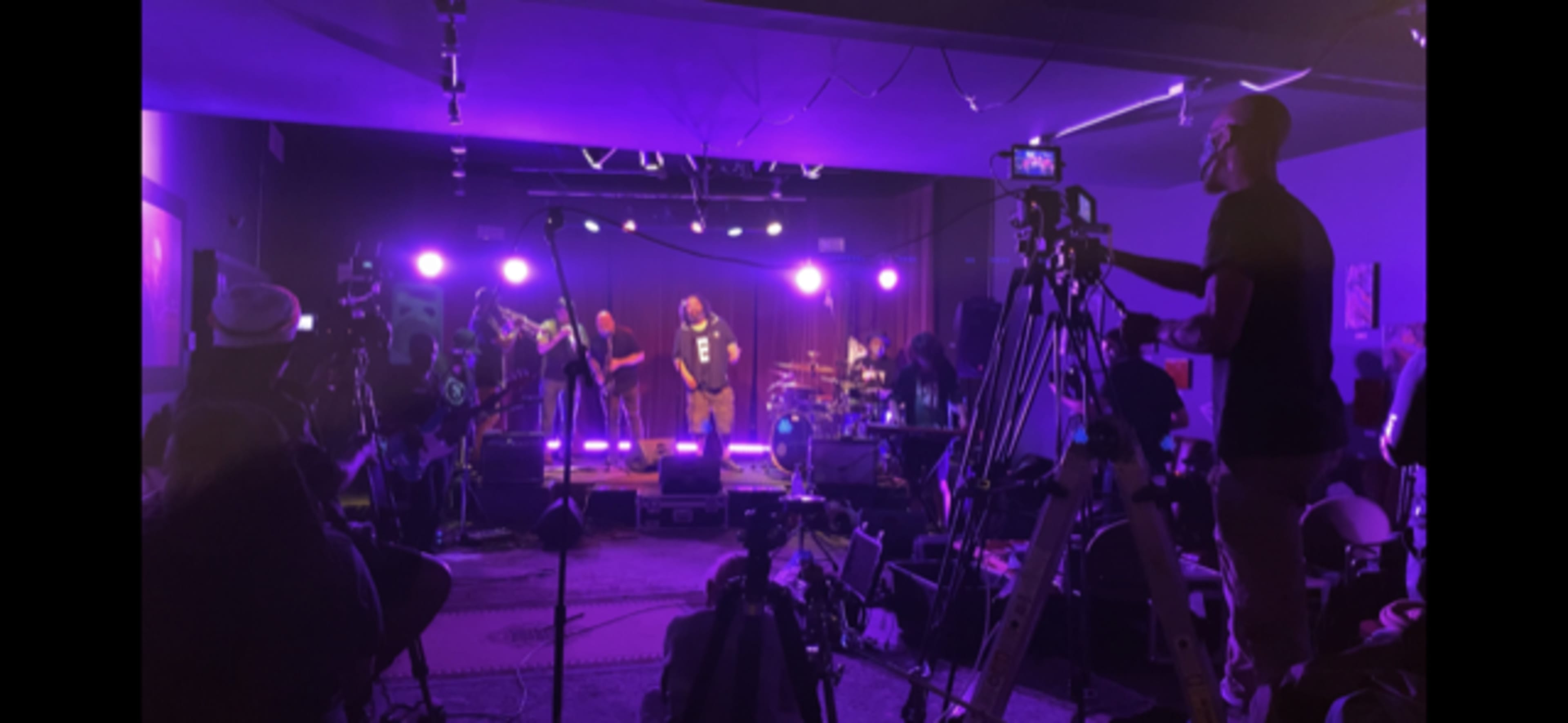 A live band performs on stage in a dimly lit venue, surrounded by cameras and crew members capturing the event.