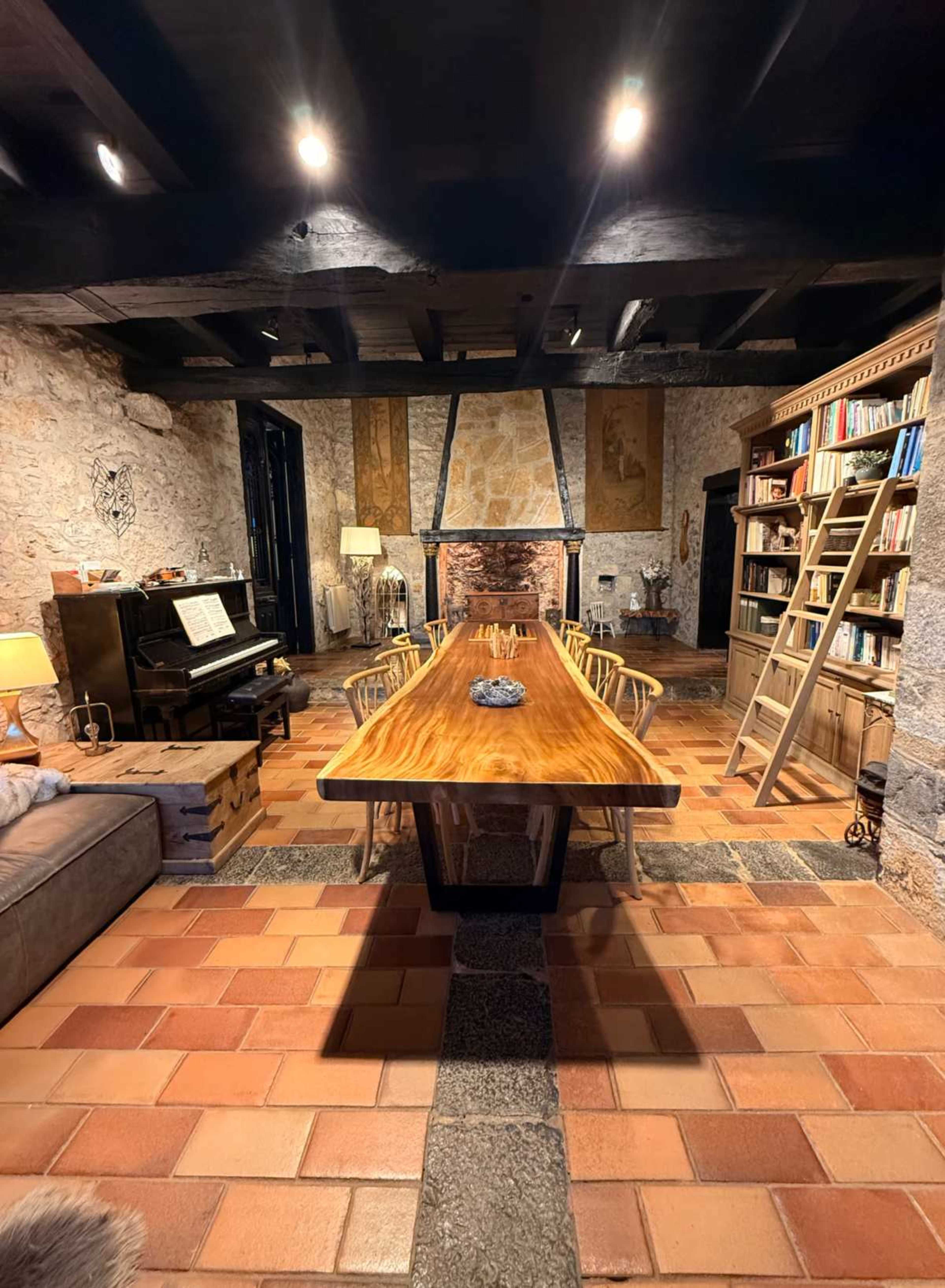 The image shows a rustic dining room featuring a long wooden table, a leather sofa, a piano, bookshelves filled with books, and stone walls.