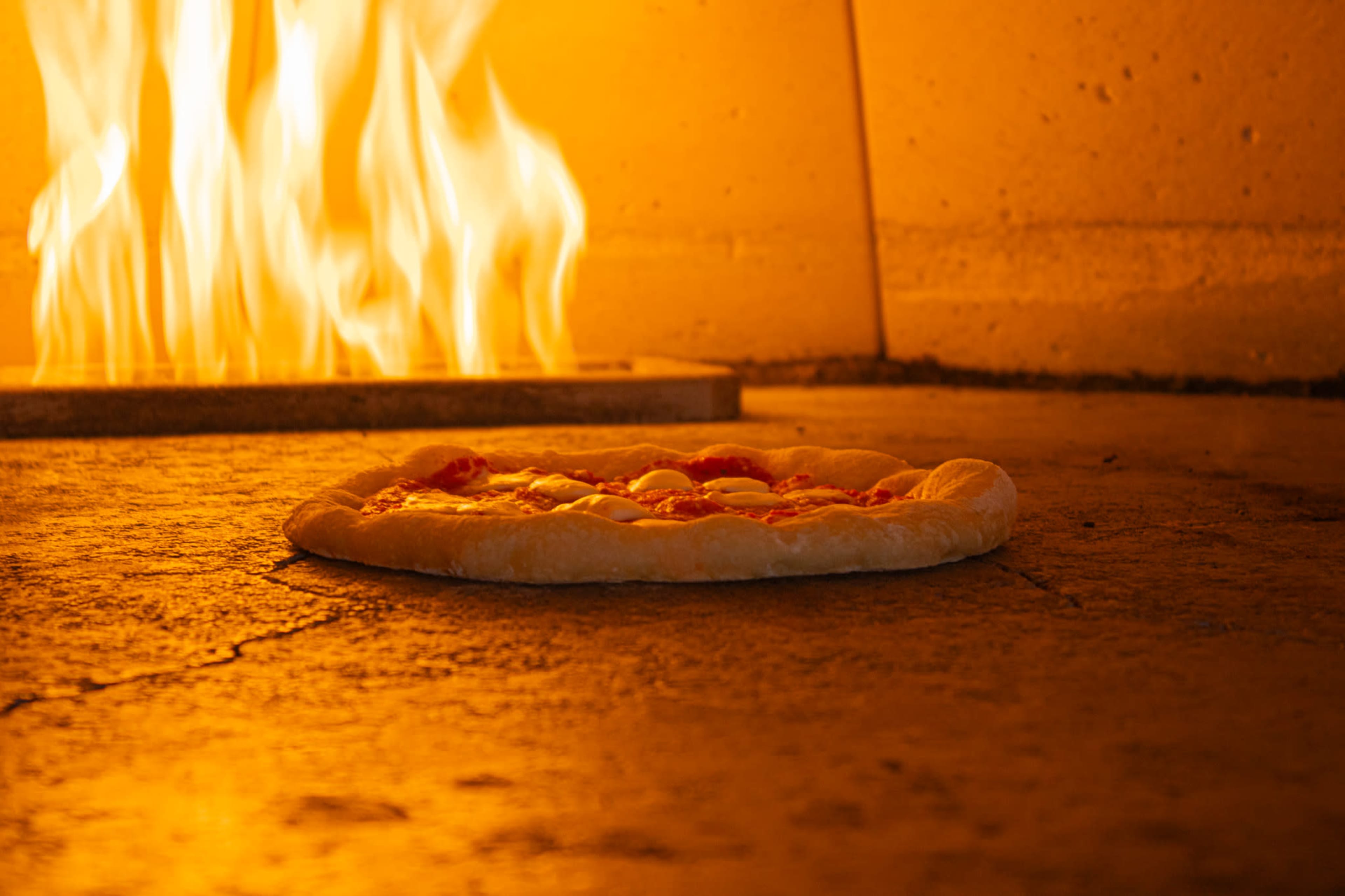 A pizza is placed on the floor of a brick oven with flames visible in the background.