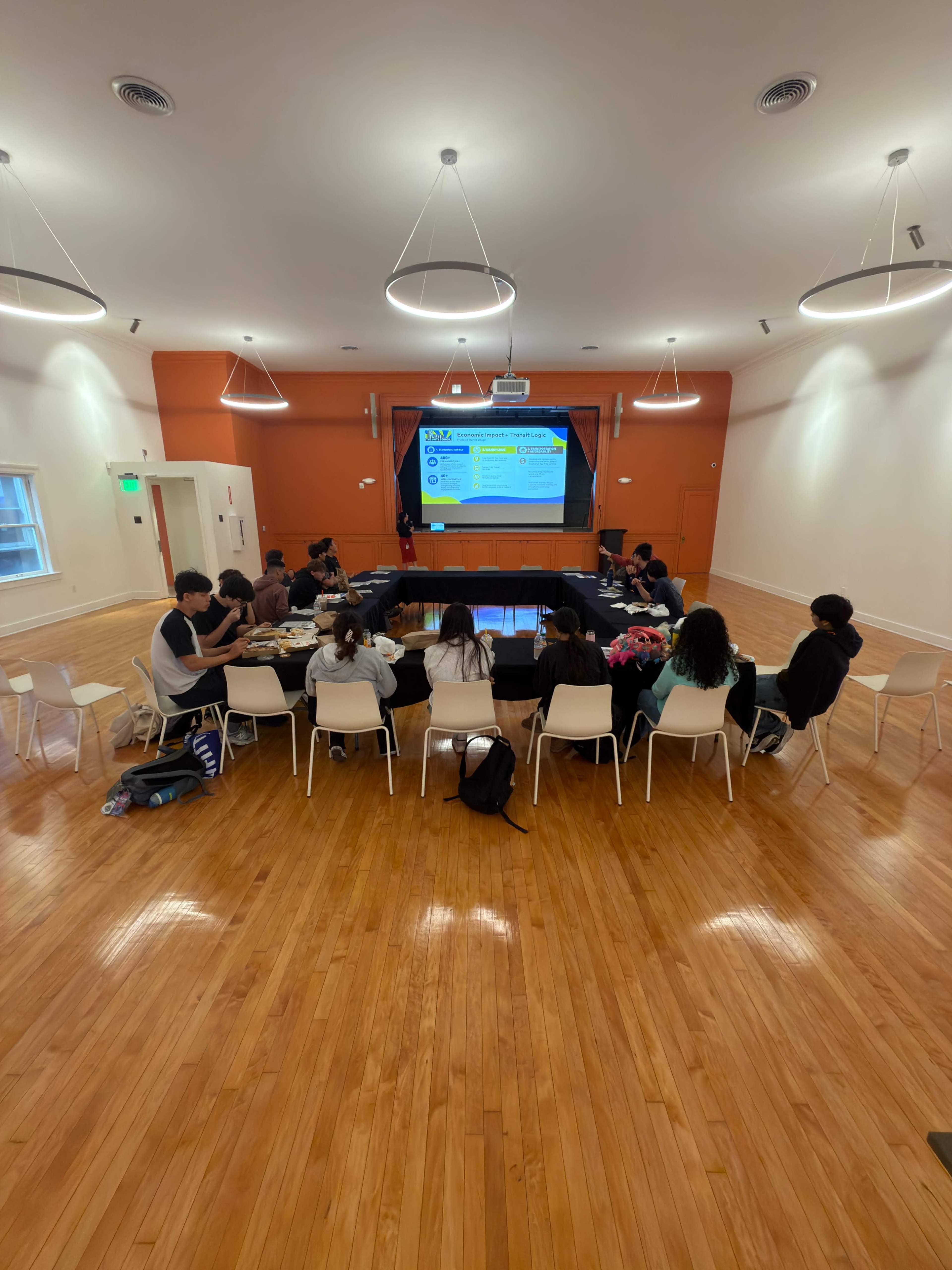 A group of people is seated in a well-lit room with wooden flooring, attentively facing a large screen displaying presentation content.