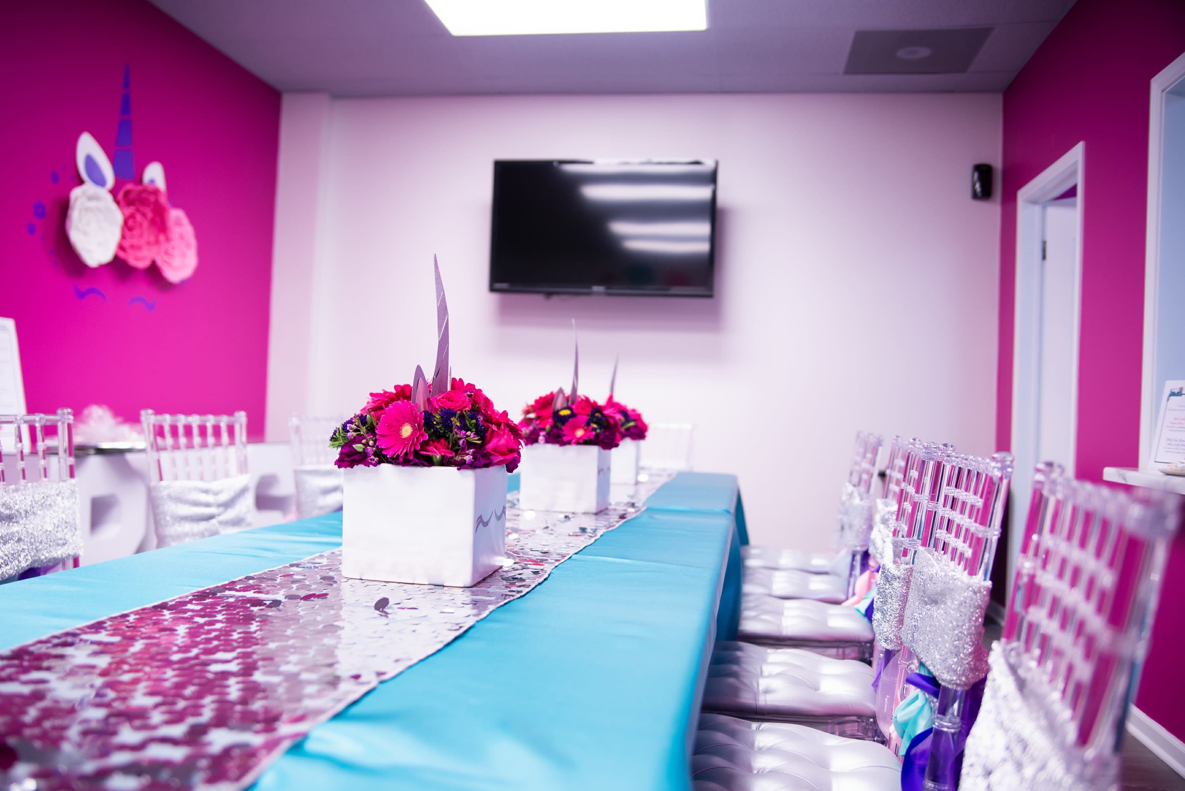 A dining area is decorated with a long table covered in turquoise cloth, adorned with a silver sequin runner and floral centerpieces, in a room with pink walls and a mounted television.