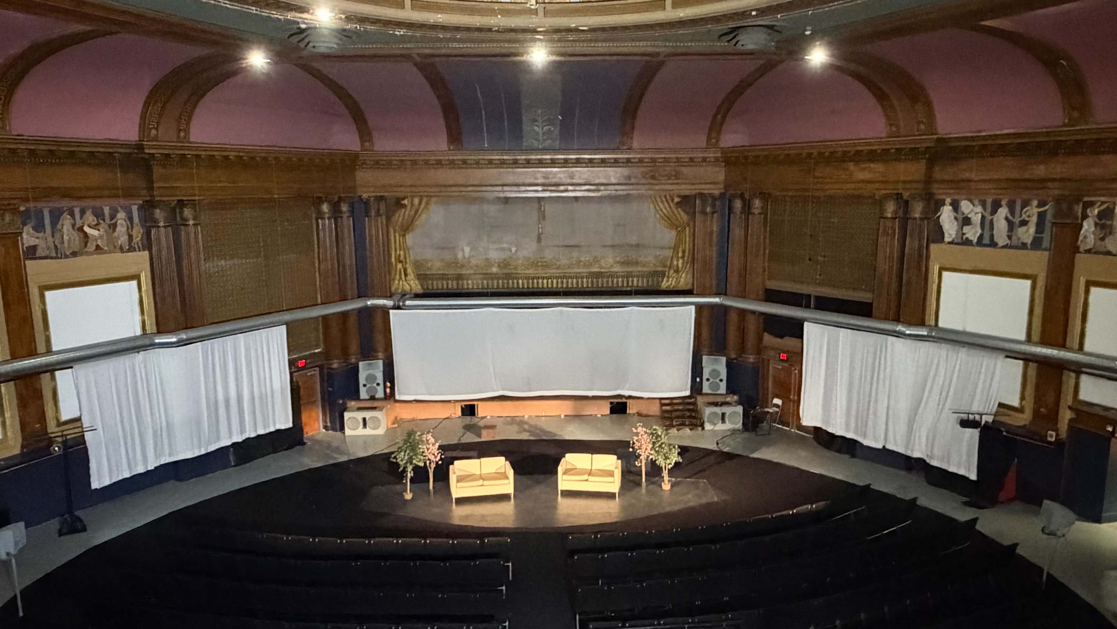 The image shows a large, empty theater with wooden walls, two chairs on a stage, and draped white curtains hanging at the sides.