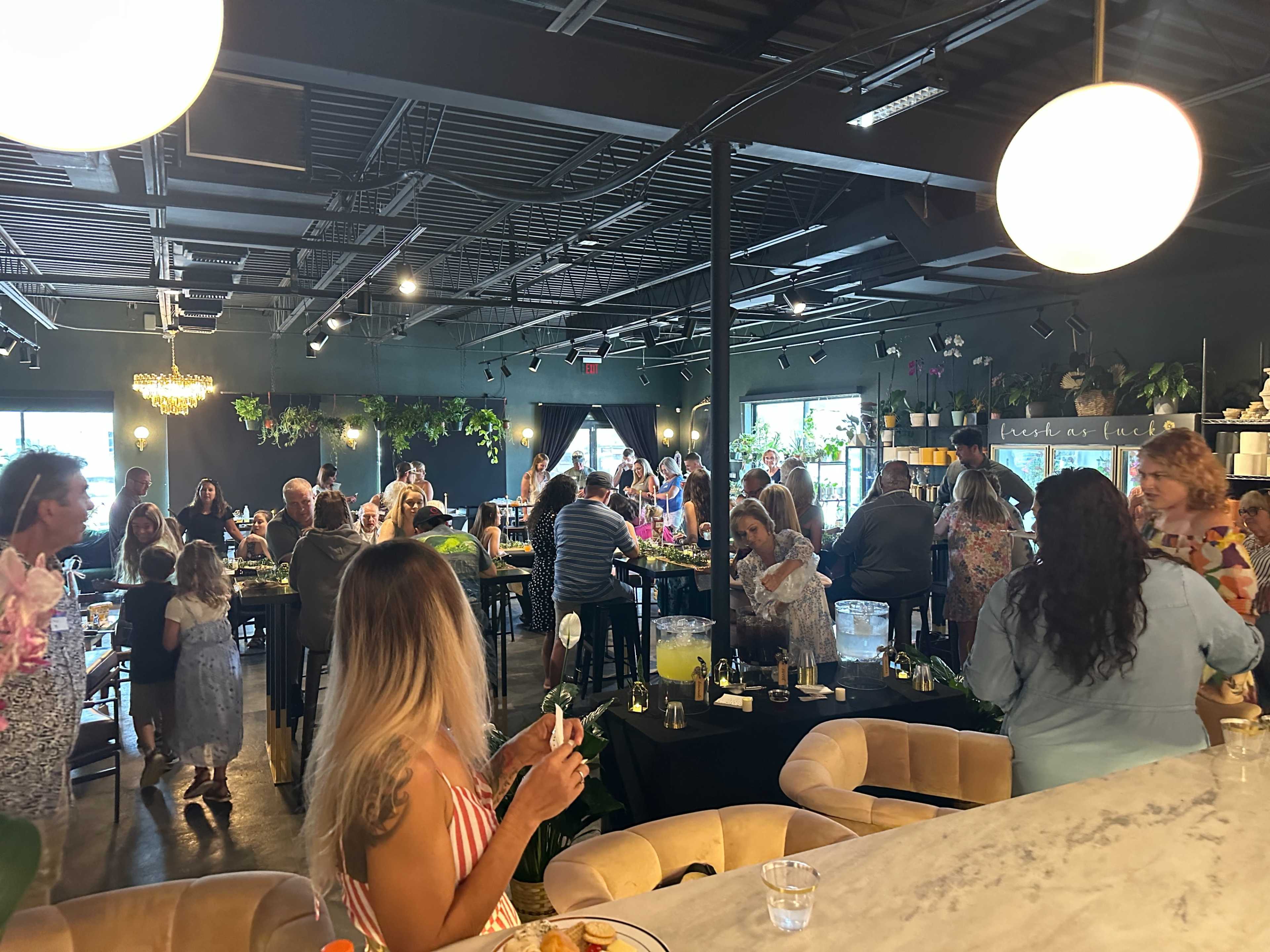 The image shows a bustling restaurant filled with people dining and engaging in conversation, with greenery and decorative lights enhancing the ambiance.