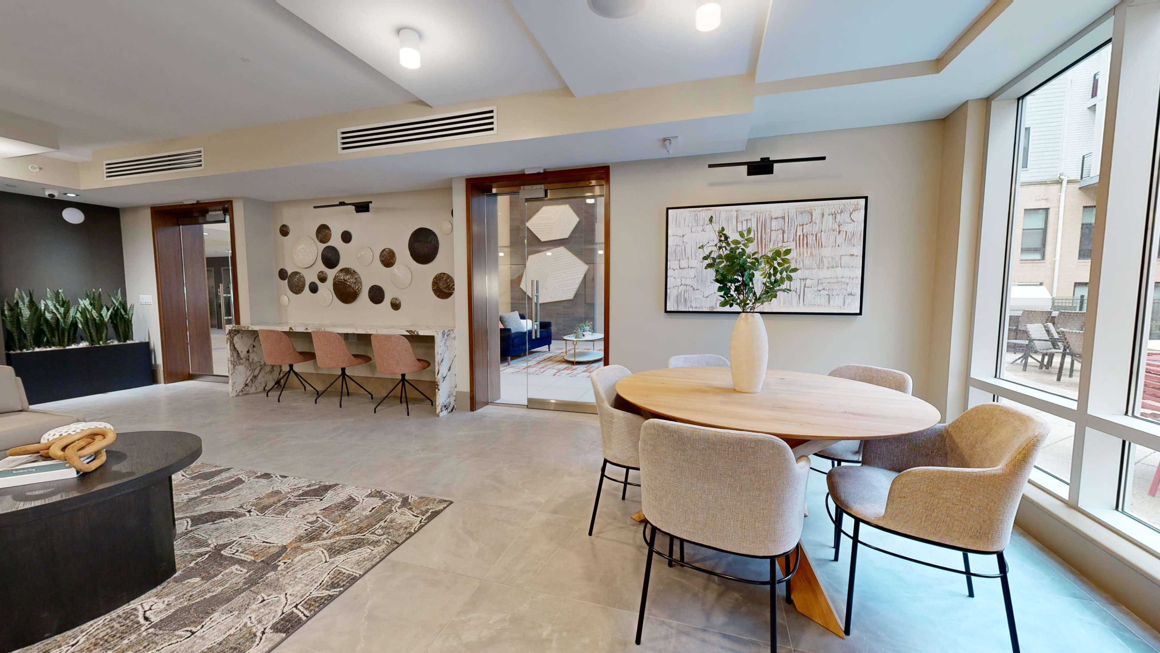 A modern interior space featuring a round dining table surrounded by upholstered chairs, a patterned rug, and decorative wall art, with large windows providing natural light.