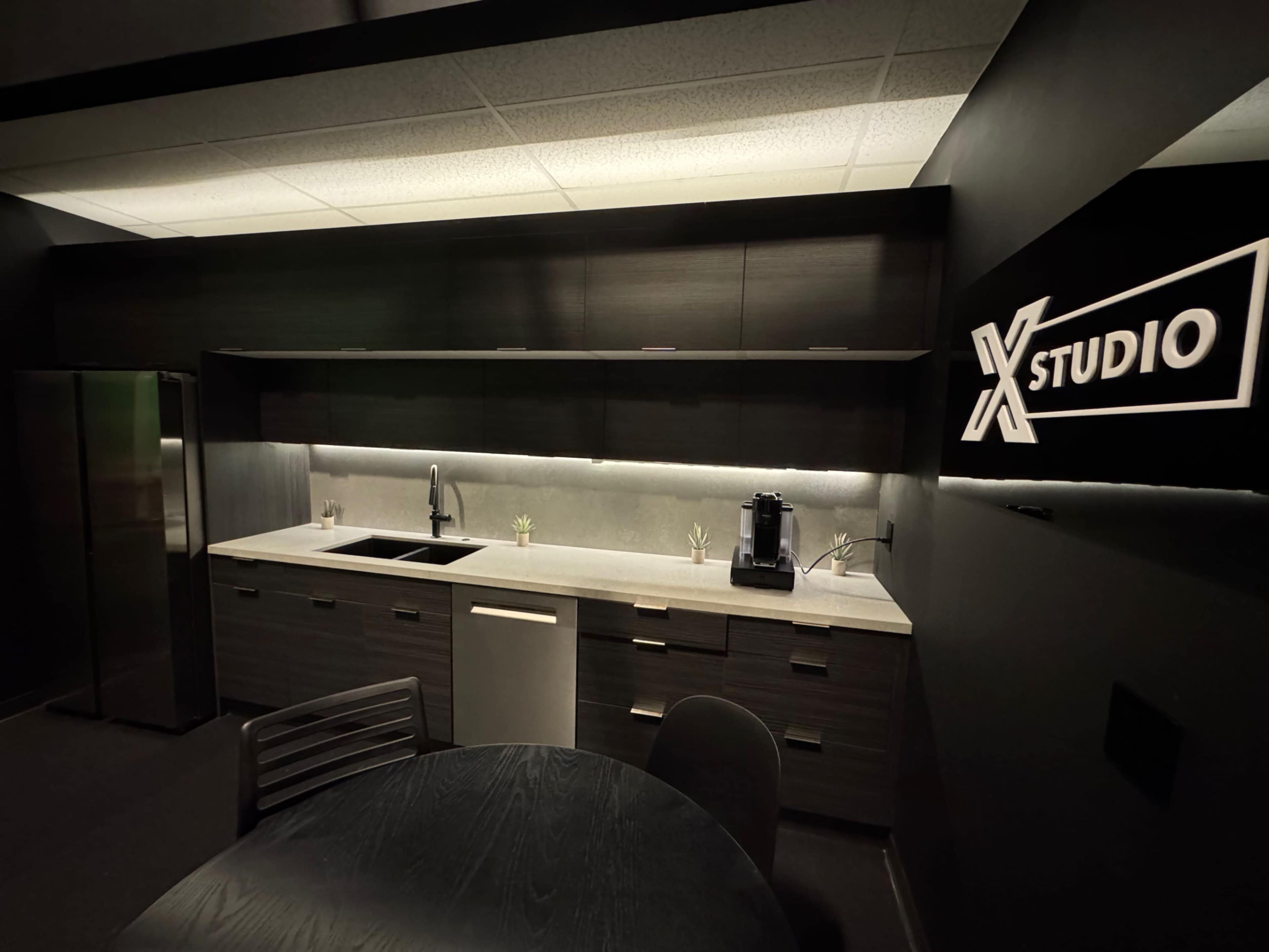 The image shows a modern kitchen space with a dark color scheme, featuring sleek cabinets, a sink, a coffee machine, and a round dining table.