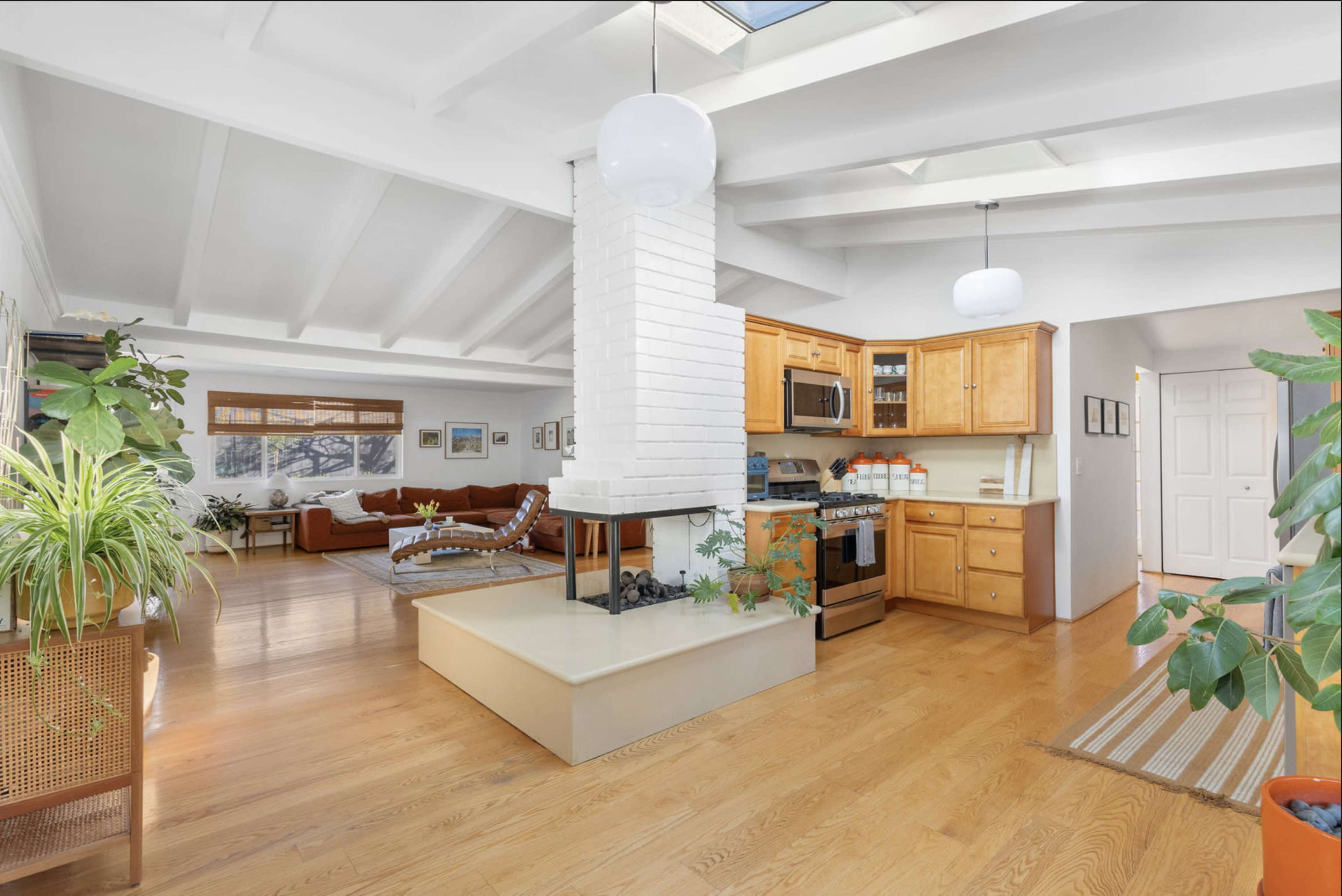 The image shows an open-concept living space with a kitchen, dining area, and living room featuring wood beams, a brick fireplace, and large windows.