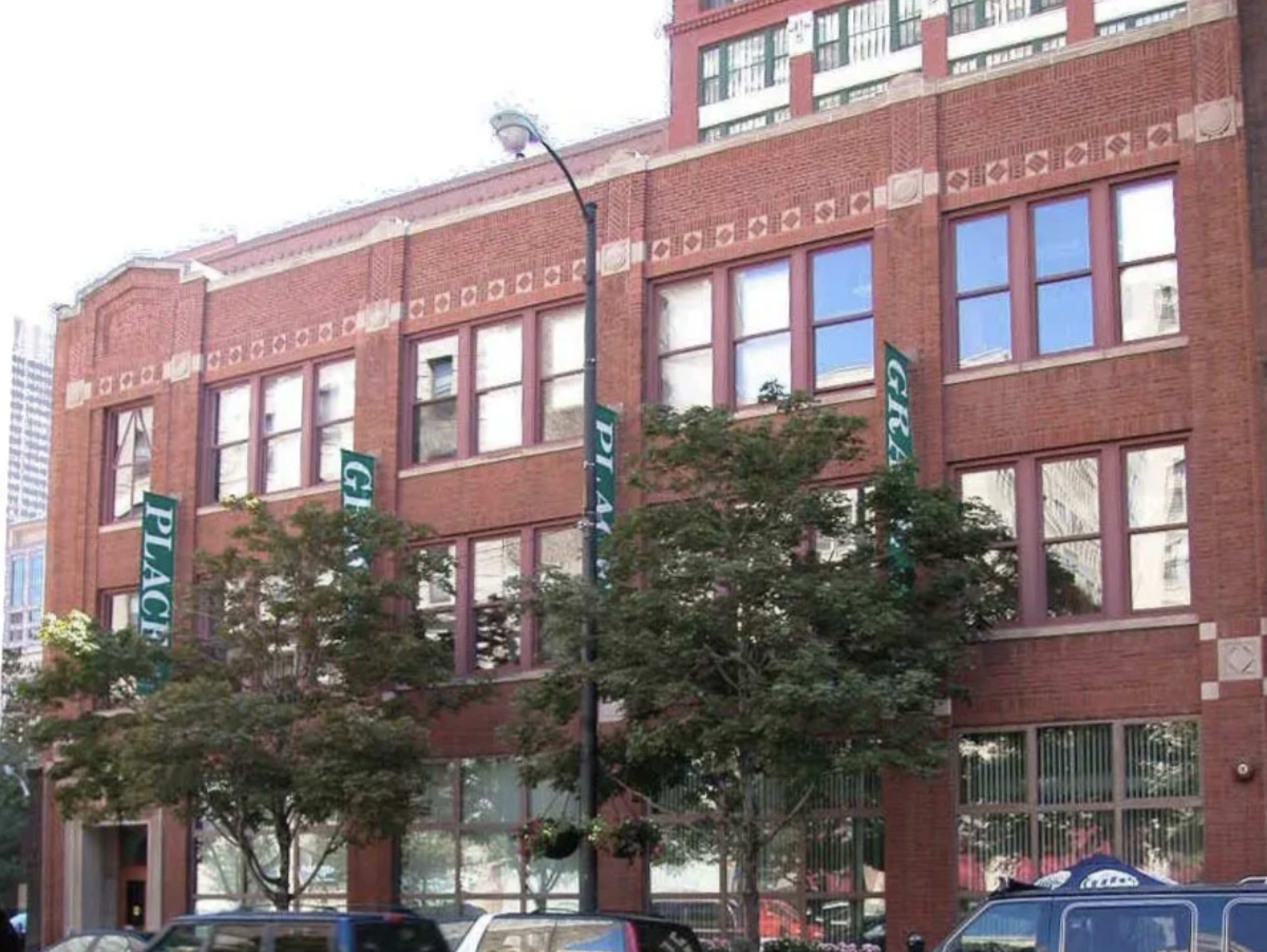 The image shows a red brick building with large windows, displaying green banners and surrounded by trees and parked cars.