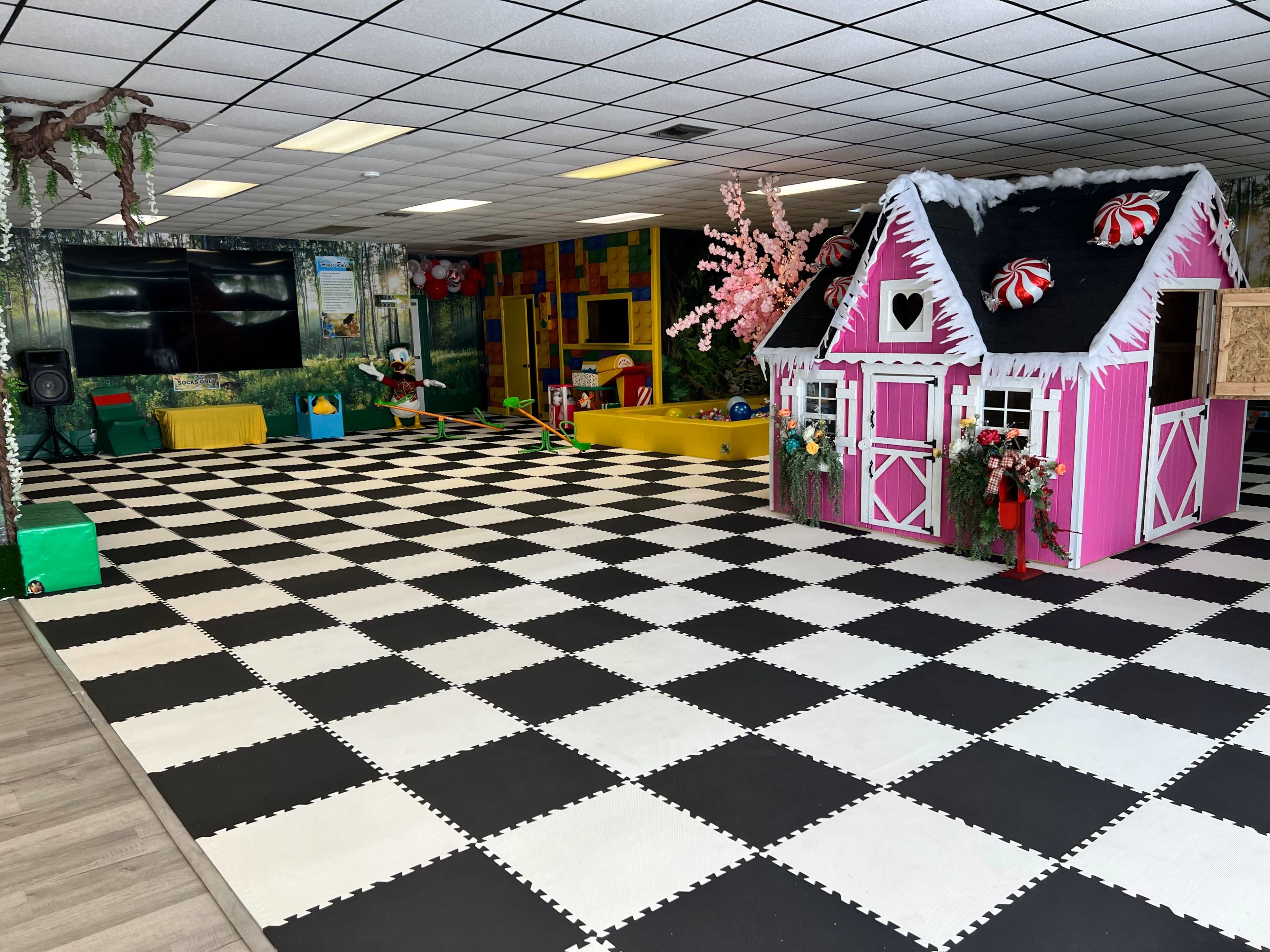 The image shows a brightly colored play space with a pink and white playhouse, black and white checkered flooring, and various play equipment.