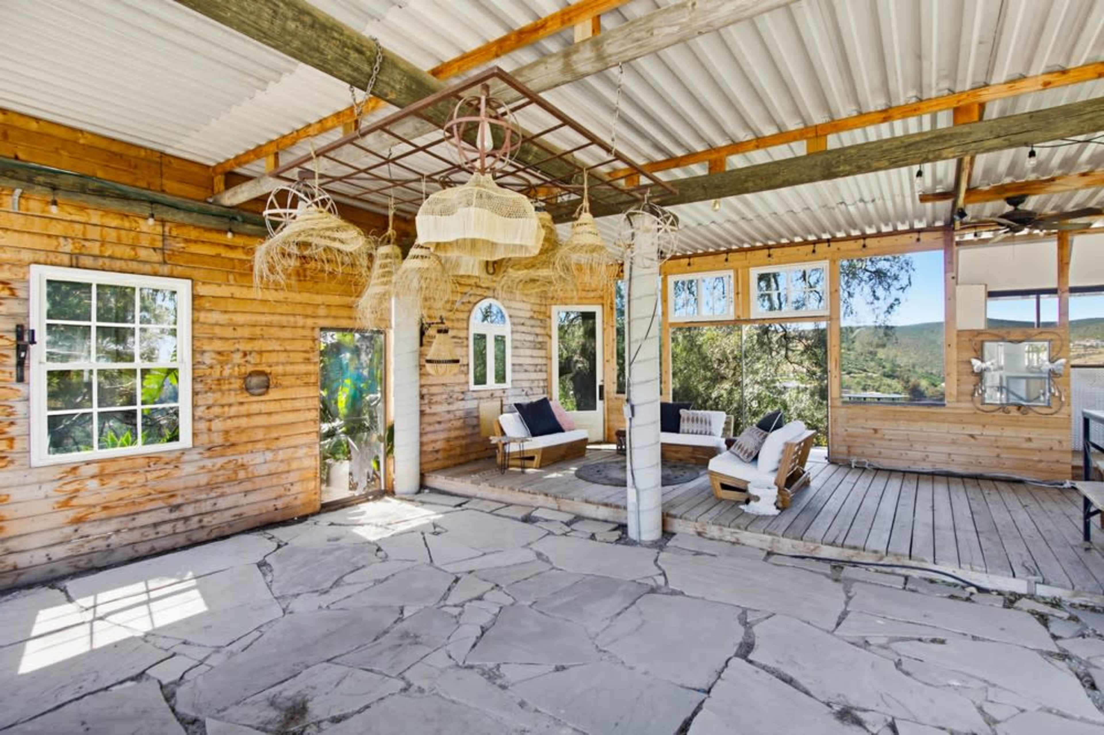 The image shows a rustic wooden interior with large windows, a seating area, and a stone floor, all illuminated by natural light.