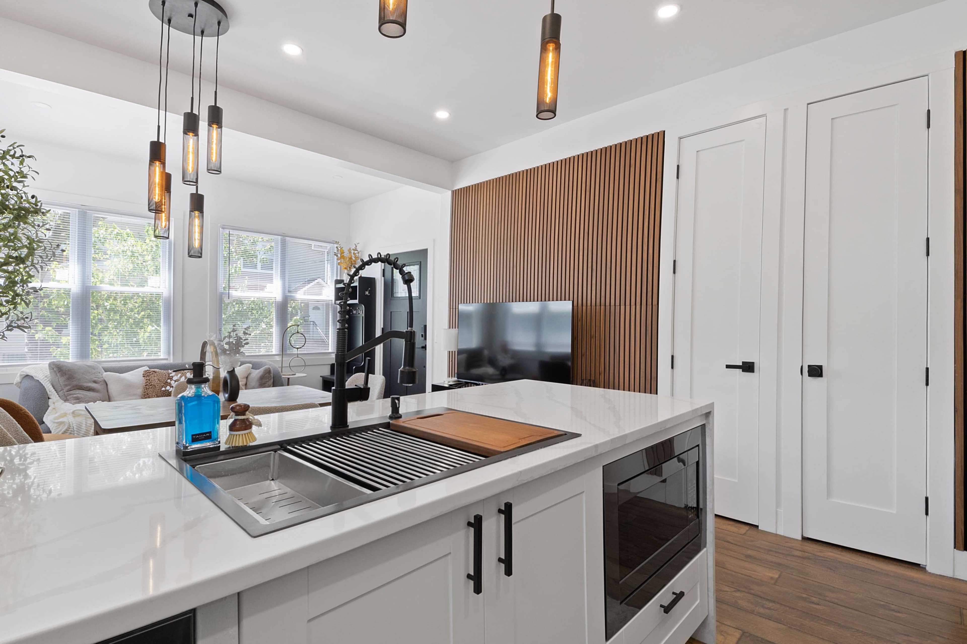 A modern kitchen featuring a white countertop with a sink, a wooden accent wall, pendant lighting, and a living area visible in the background.