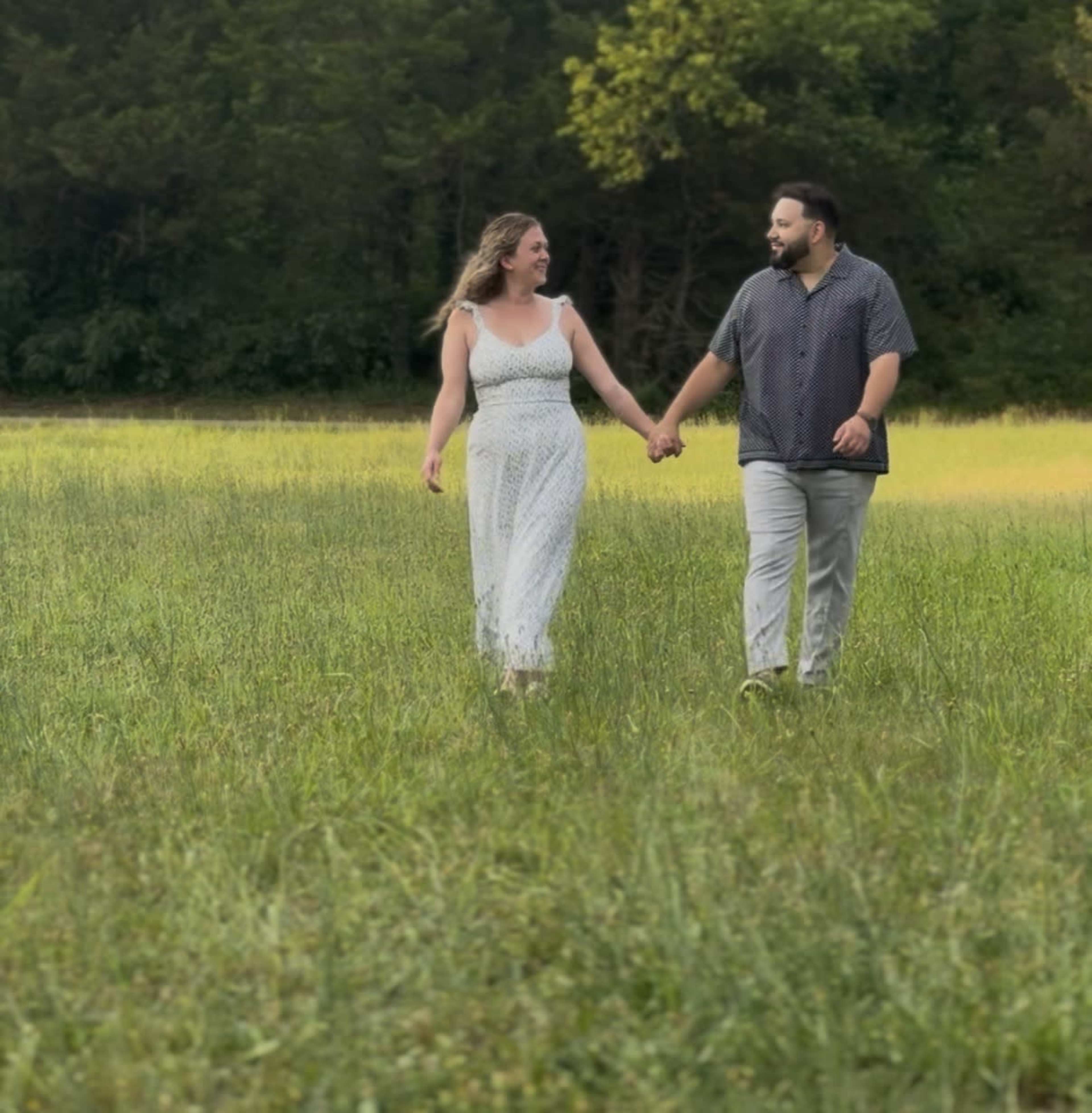 A couple walks hand in hand through a grassy field under a clear sky.
