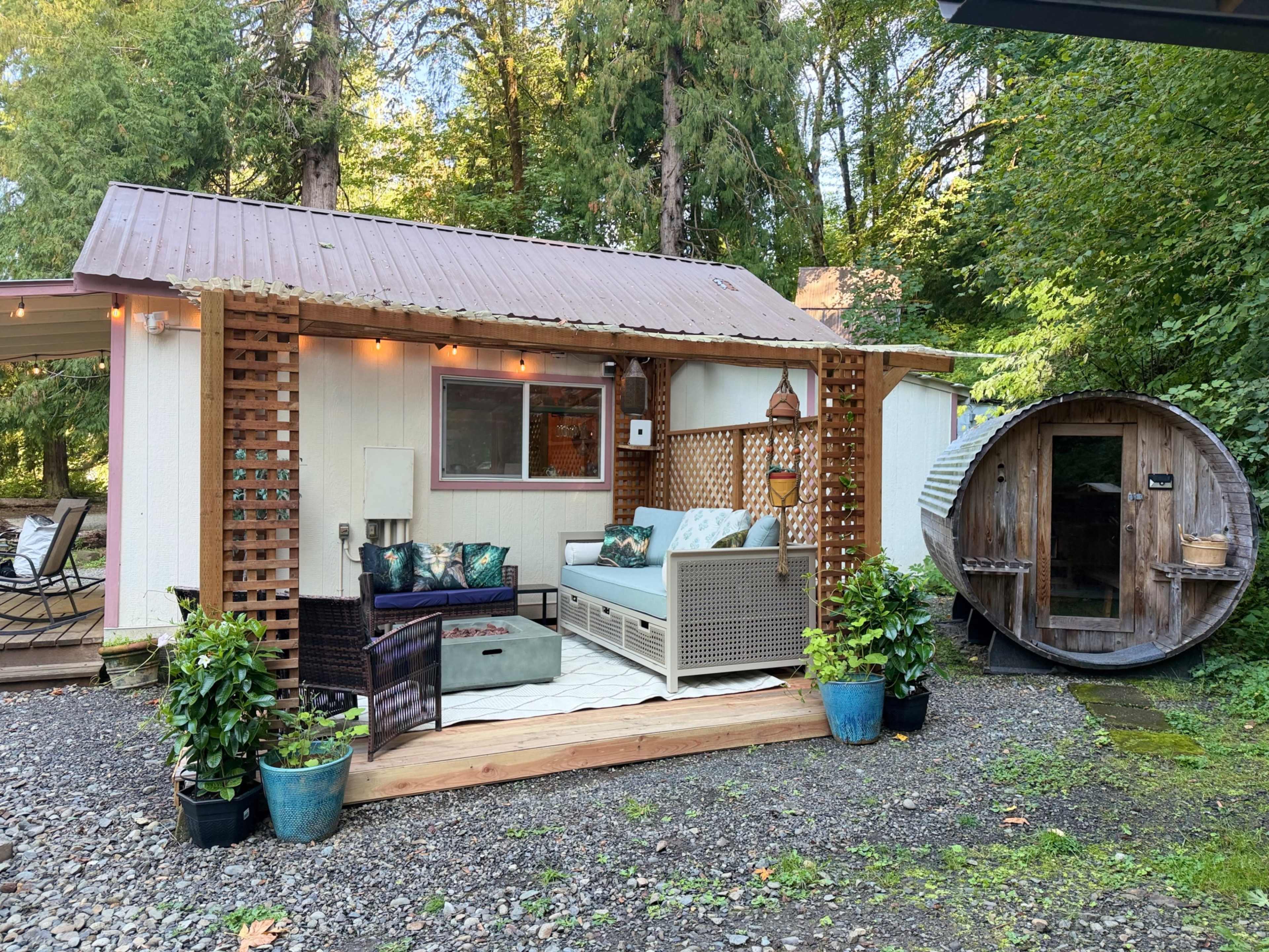 The image shows a small, rustic cabin with a wooden deck, outdoor seating area, and a round wooden sauna nearby, surrounded by greenery.