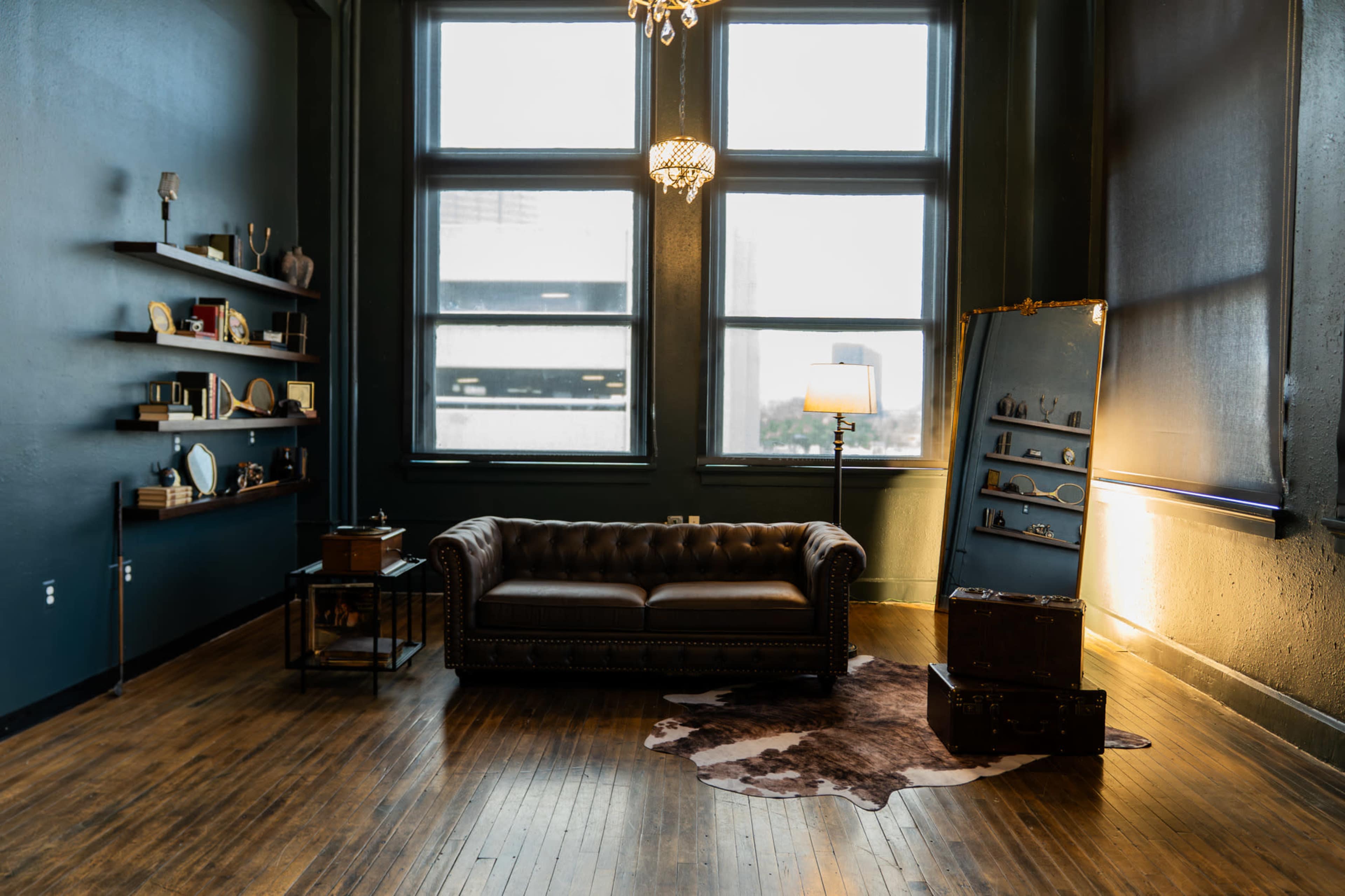 The image shows a cozy interior space featuring a leather couch, a wooden floor, large windows with natural light, and various decorative items on shelves and a display stand.